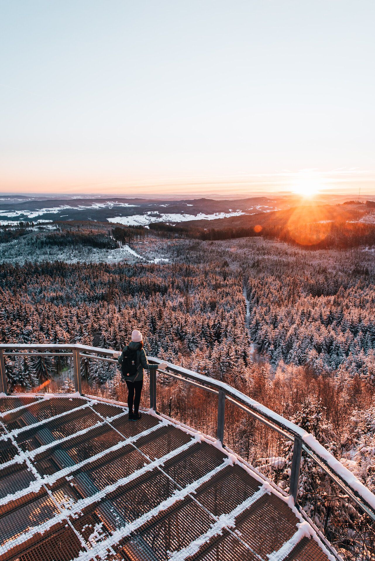 Die winterliche Landschaft strahlt in sanften Pastelltönen, während die Sonne langsam am Horizont aufgeht. Ein Wanderer genießt die frische, klare Luft und den atemberaubenden Blick auf die schneebedeckten Wälder, die in der Morgensonne glitzern.