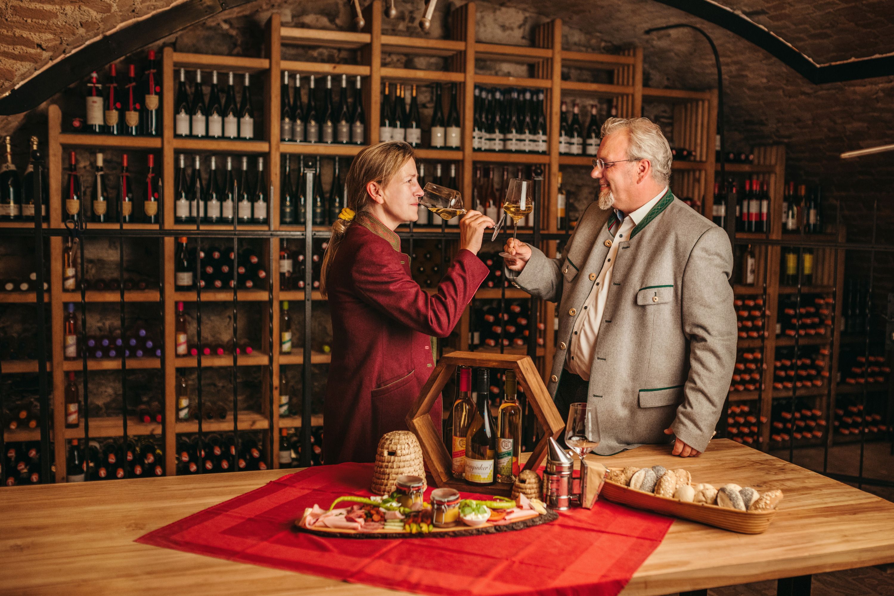 Zwei Personen stoßen mit Weingläsern in einem Weinkeller an, umgeben von Weinflaschen und einem Tisch mit Snacks.