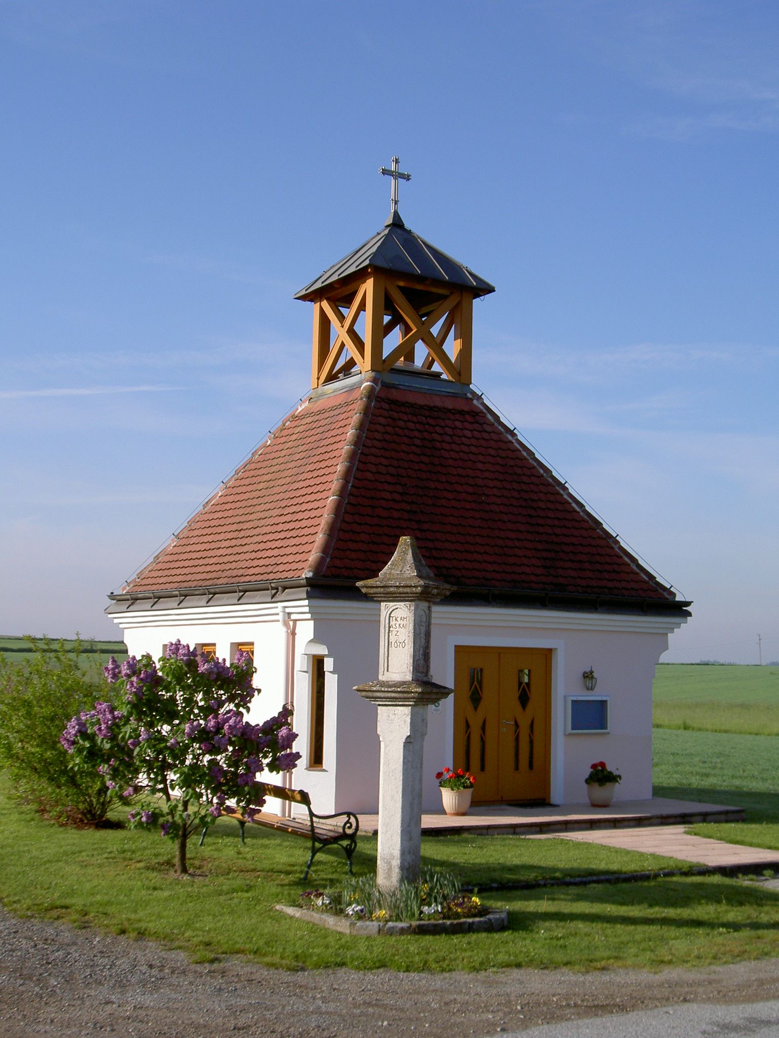 Kleine Kapelle mit rotem Dach und Kreuz, umgeben von Blumen und einer Bank, bei klarem Himmel.