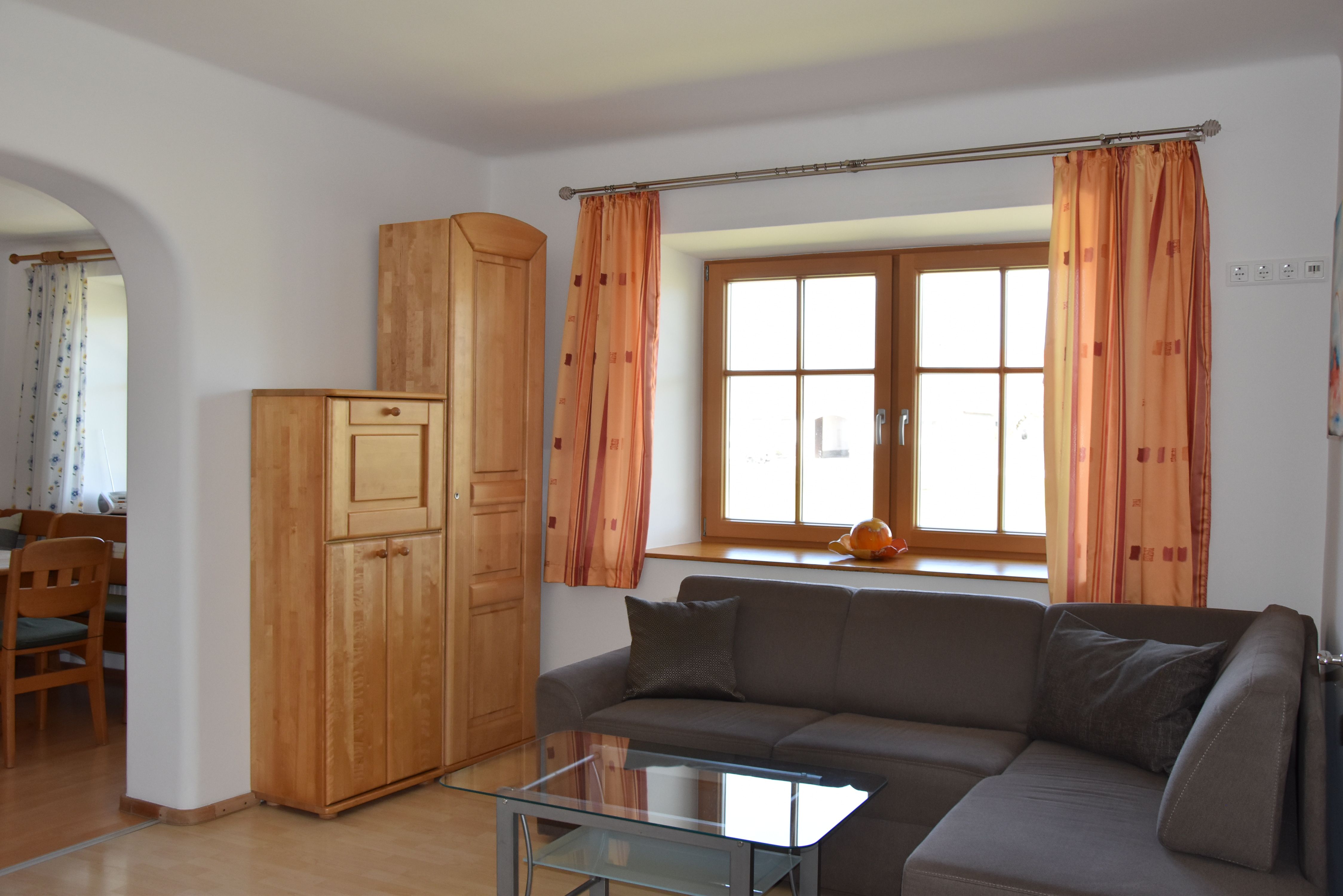 Wohnzimmer mit grauem Ecksofa, Glastisch, Schrank und Fenster mit orangefarbenen Vorhängen.