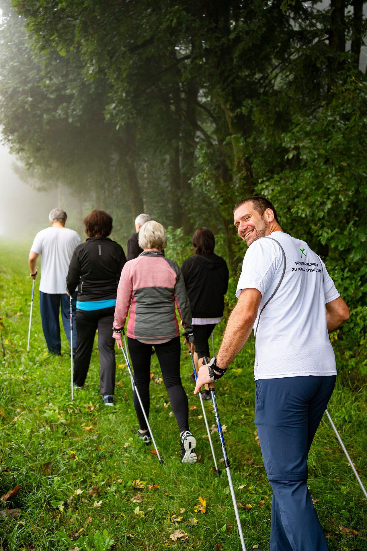 Inmitten der sanften Hügel und der frischen Luft genießen die Teilnehmer eine belebende Nordic Walking-Tour. Die dichten Bäume spenden Schatten und die Nebelschwaden verleihen der Szenerie eine mystische Atmosphäre. Hier wird Gesundheit und Wohlbefinden in der Natur großgeschrieben.