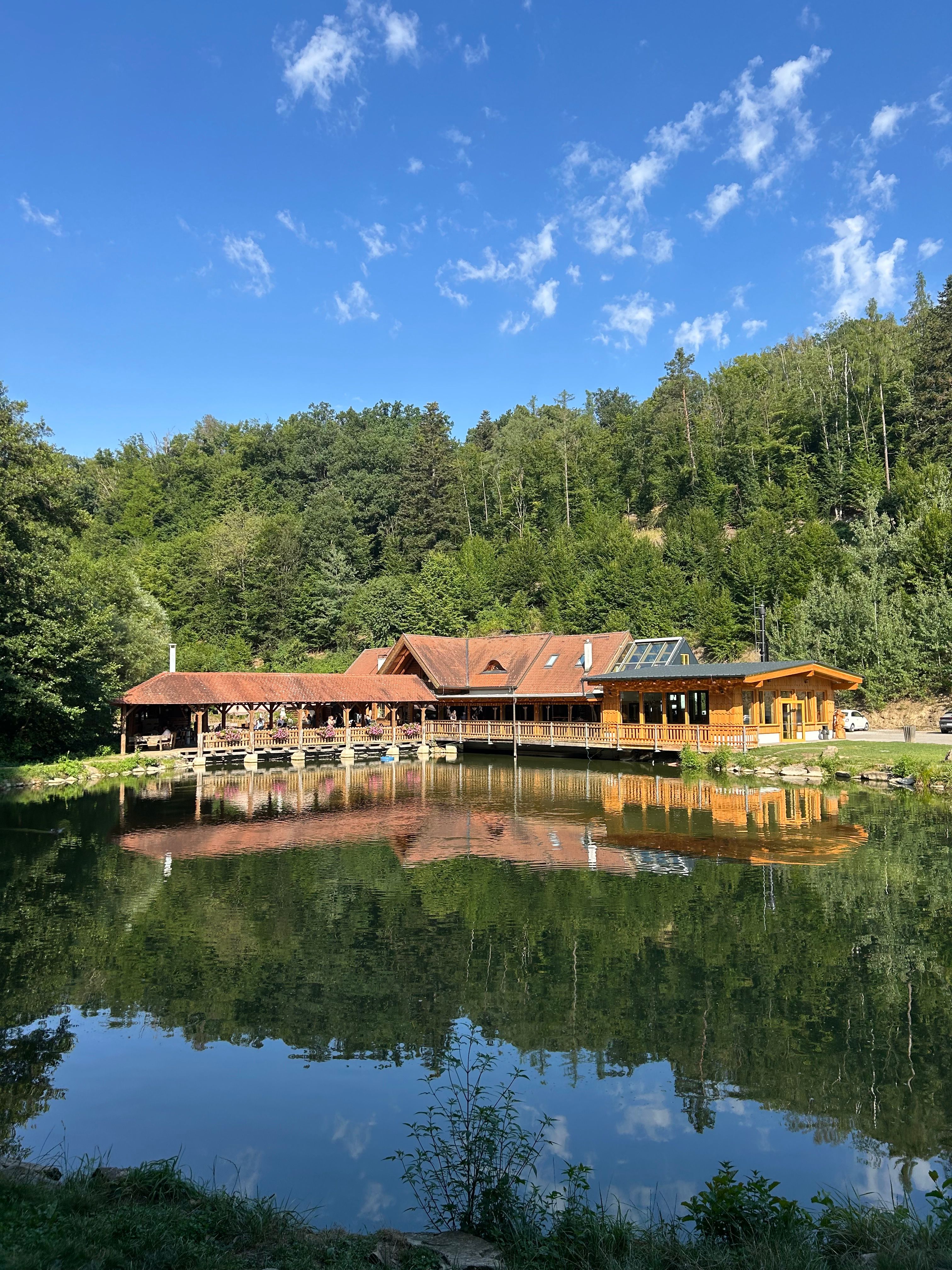 Ein Holzgebäude am Ufer eines Sees, umgeben von Wald, spiegelt sich im Wasser. Der Himmel ist blau mit wenigen Wolken.