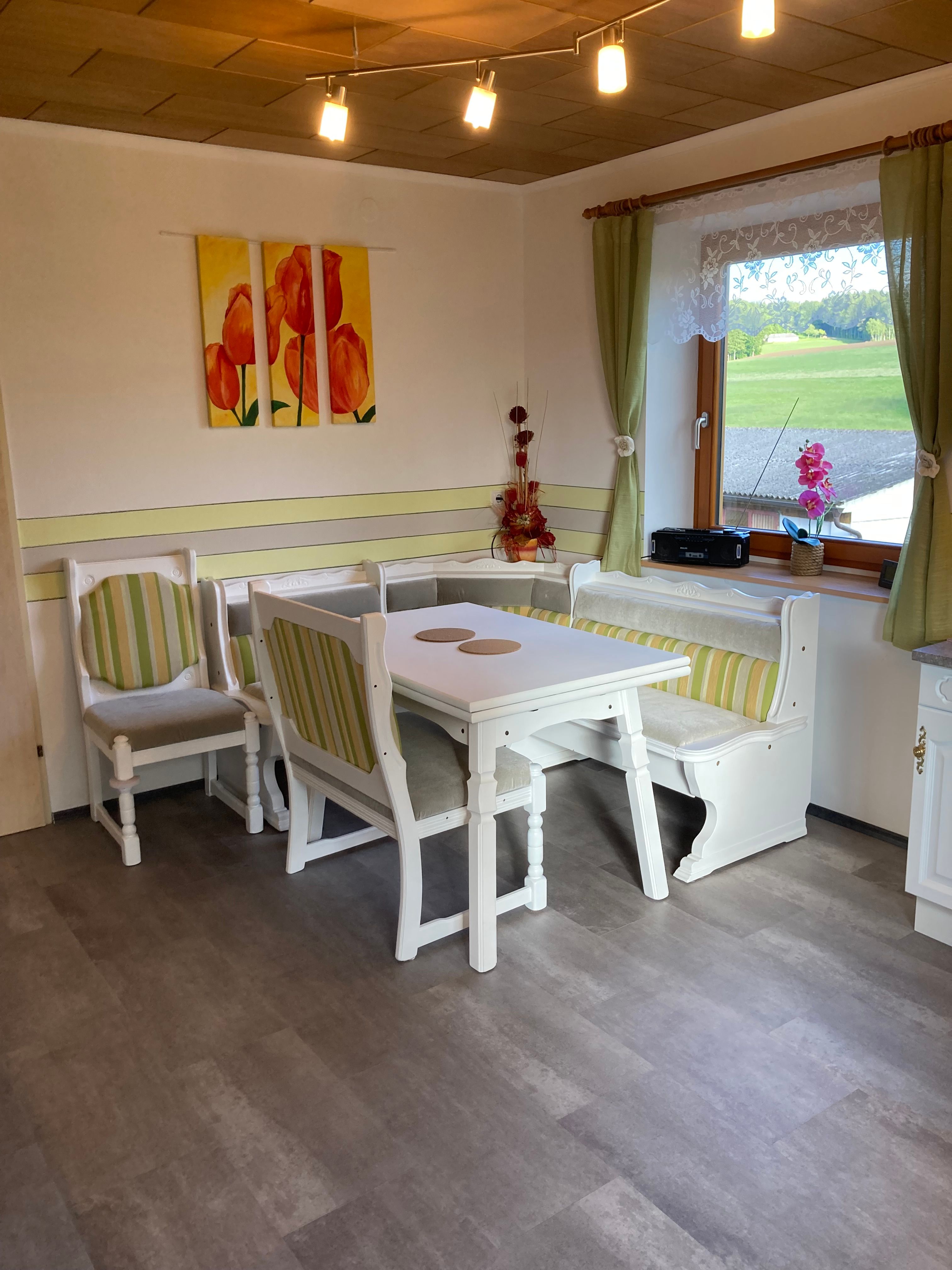 Gemütliche Essecke mit Holzmöbeln und grün-weiß gestreiften Polstern, Fenster mit Blick ins Grüne, Tulpenbild an der Wand.