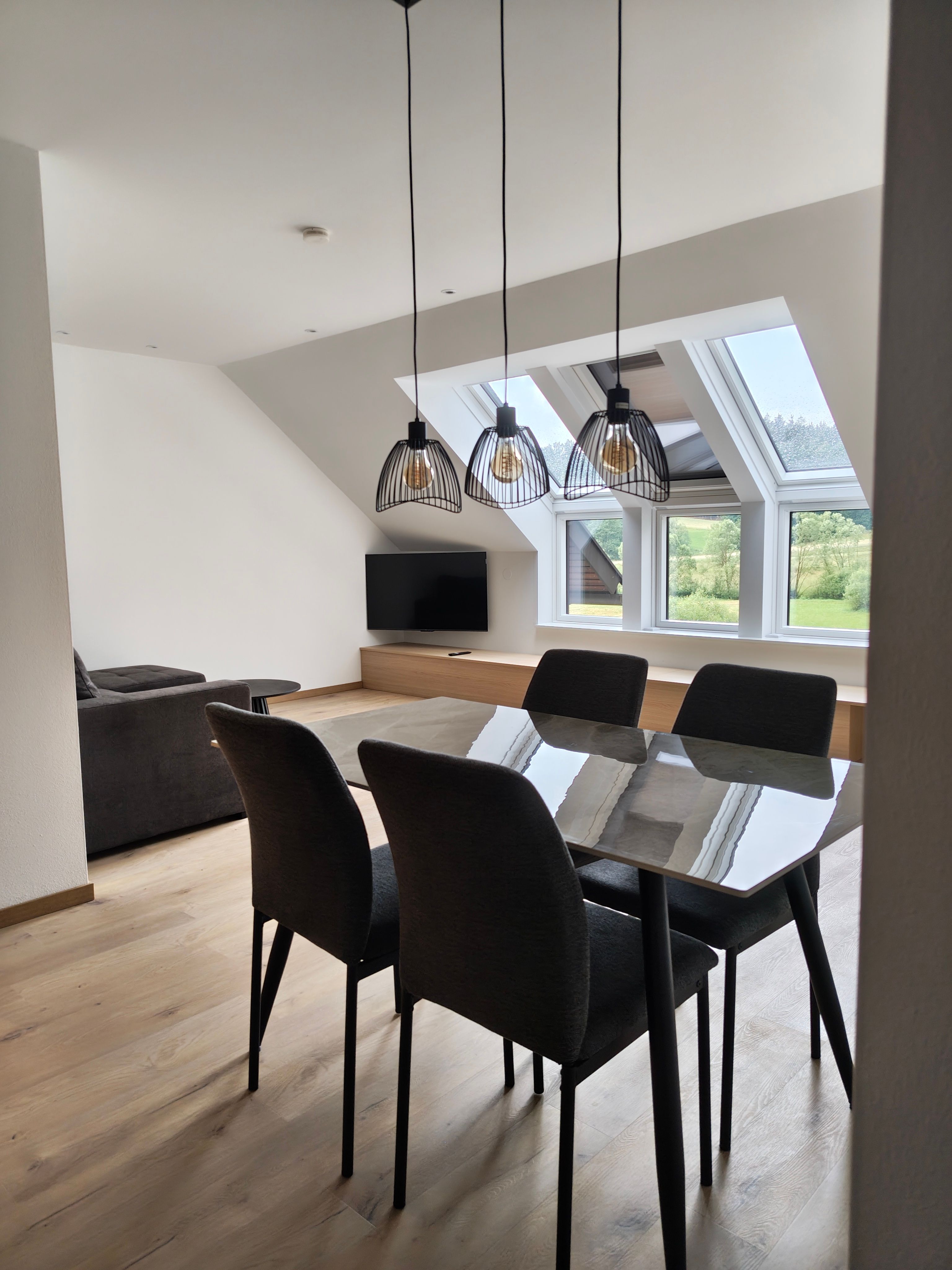 Modernes Esszimmer mit Glas-Esstisch, vier dunklen Stühlen, drei Pendelleuchten und Blick durch Dachfenster ins Grüne.