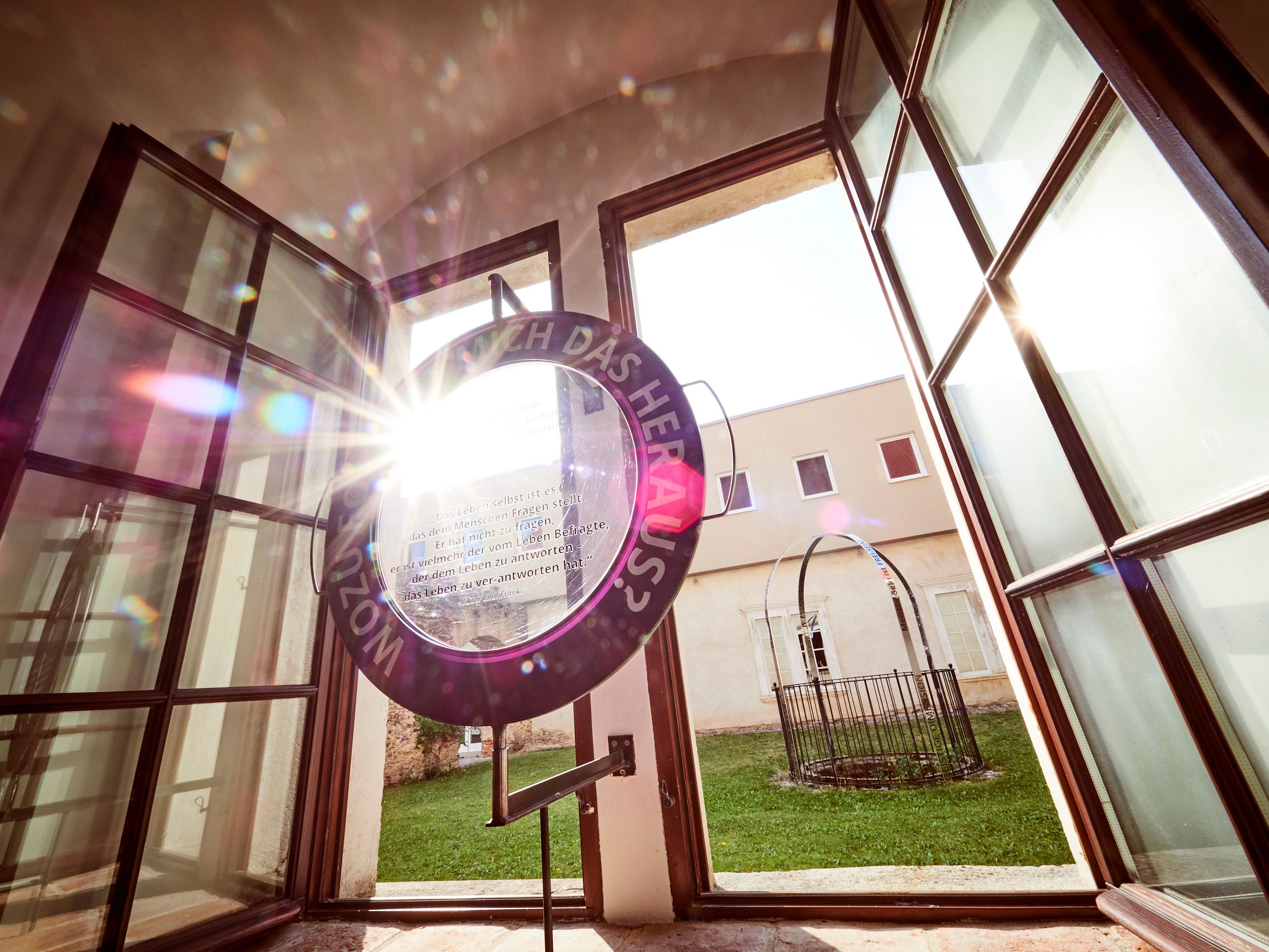 Ein offenes Fenster mit einem runden Kunstwerk, durch das Sonnenlicht strahlt, und einem Blick auf einen grünen Innenhof.