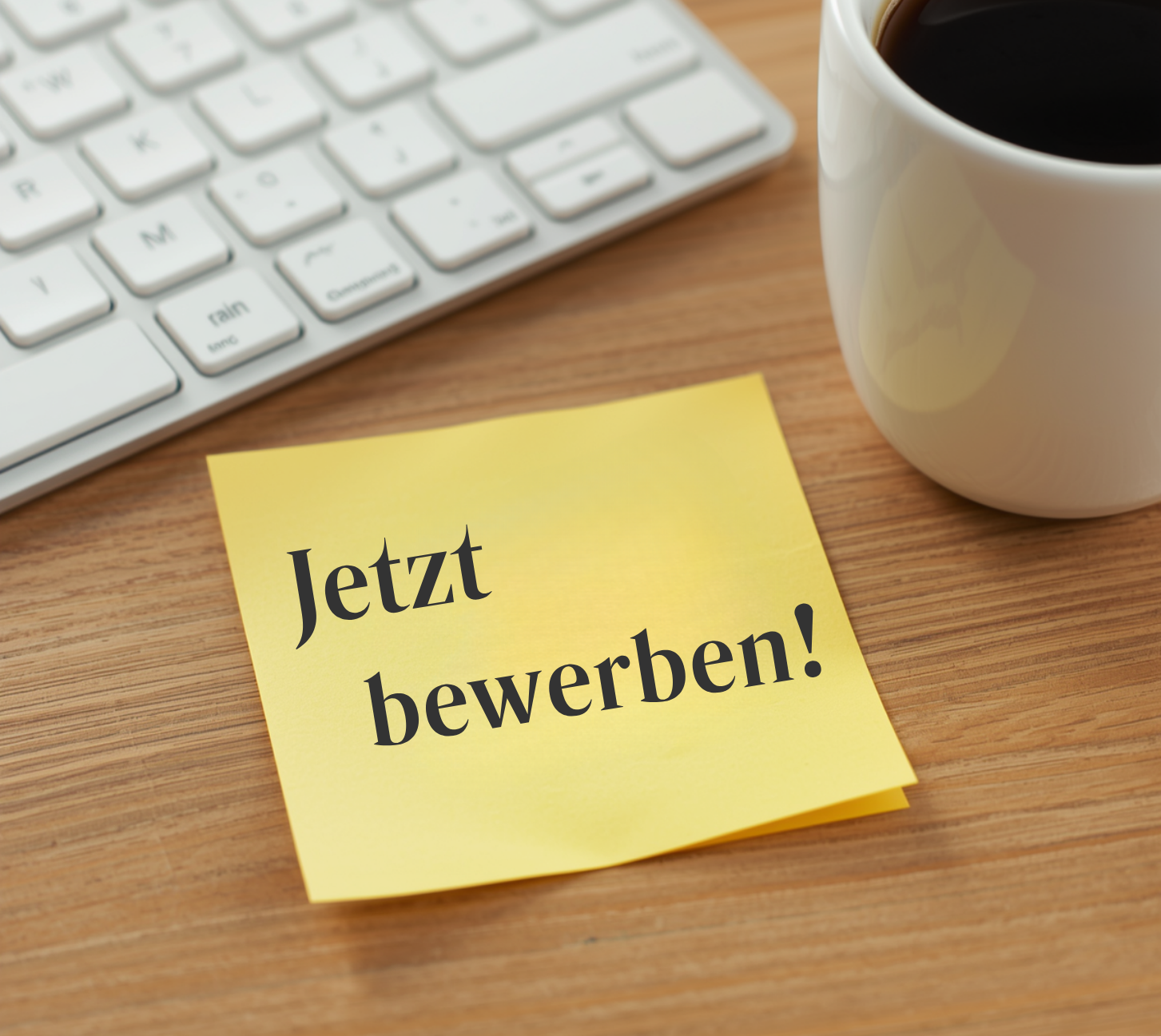 Ein Schreibtisch mit einem gelben Post-It wo "Jetzt bewerben"! steht. Im Hintergrund eine Tasse Kaffee und eine weiße Tastatur. 