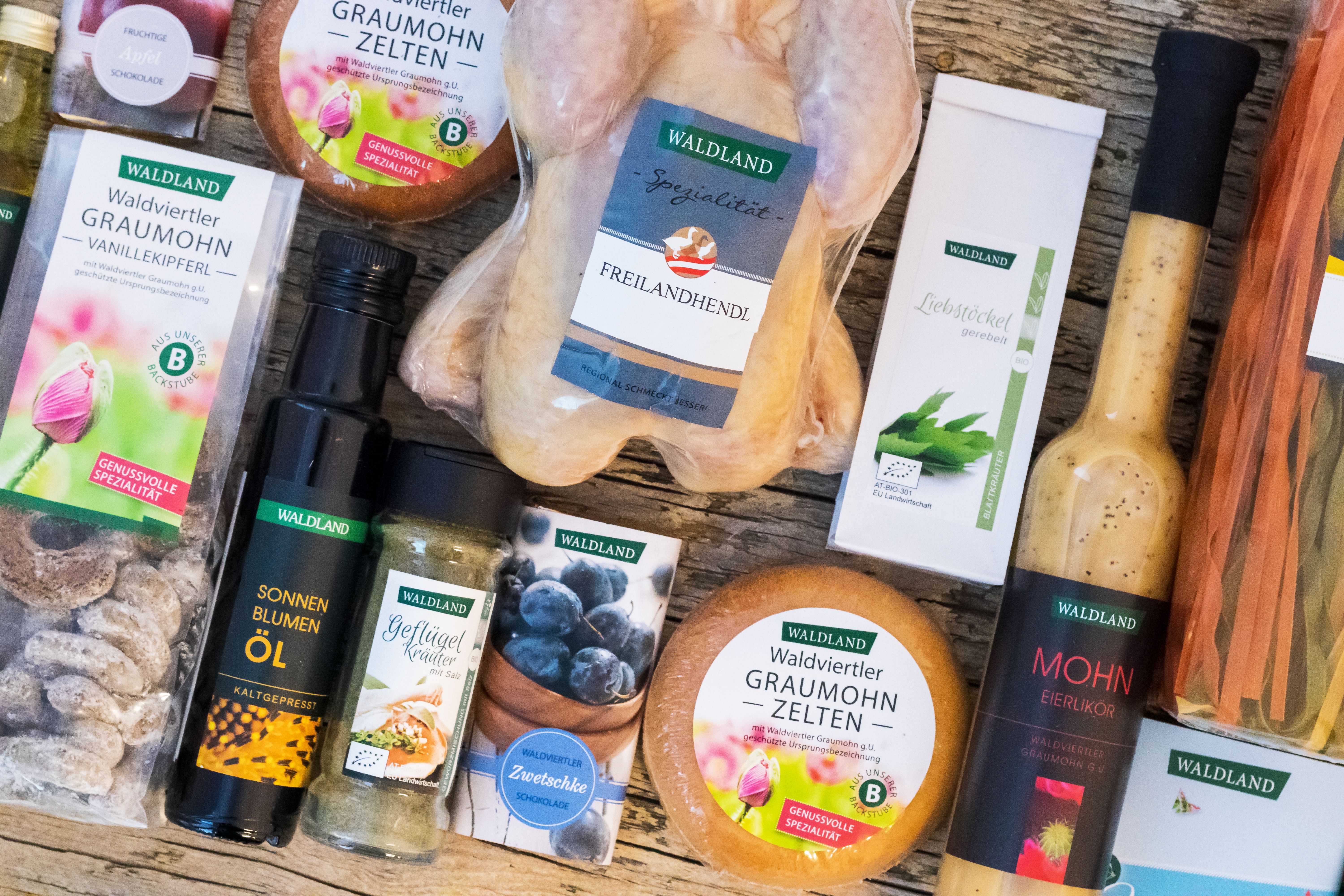 Verschiedene regionale Waldland-Produkte wie Freilandhendl, Graumohn-Zelten, Sonnenblumenöl, Gewürze, Schokolade und Likör auf einem Holztisch.