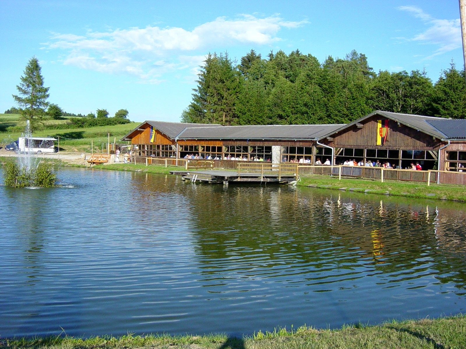 Ein Holzgebäude an einem Teich mit Springbrunnen, umgeben von Bäumen und Wiesen.