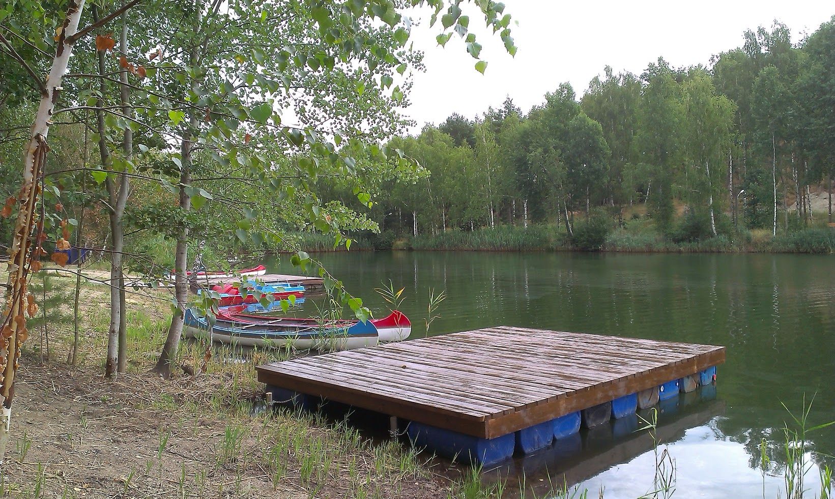 Ein ruhiger See mit einem Holzsteg und mehreren Kanus am Ufer, umgeben von Bäumen.