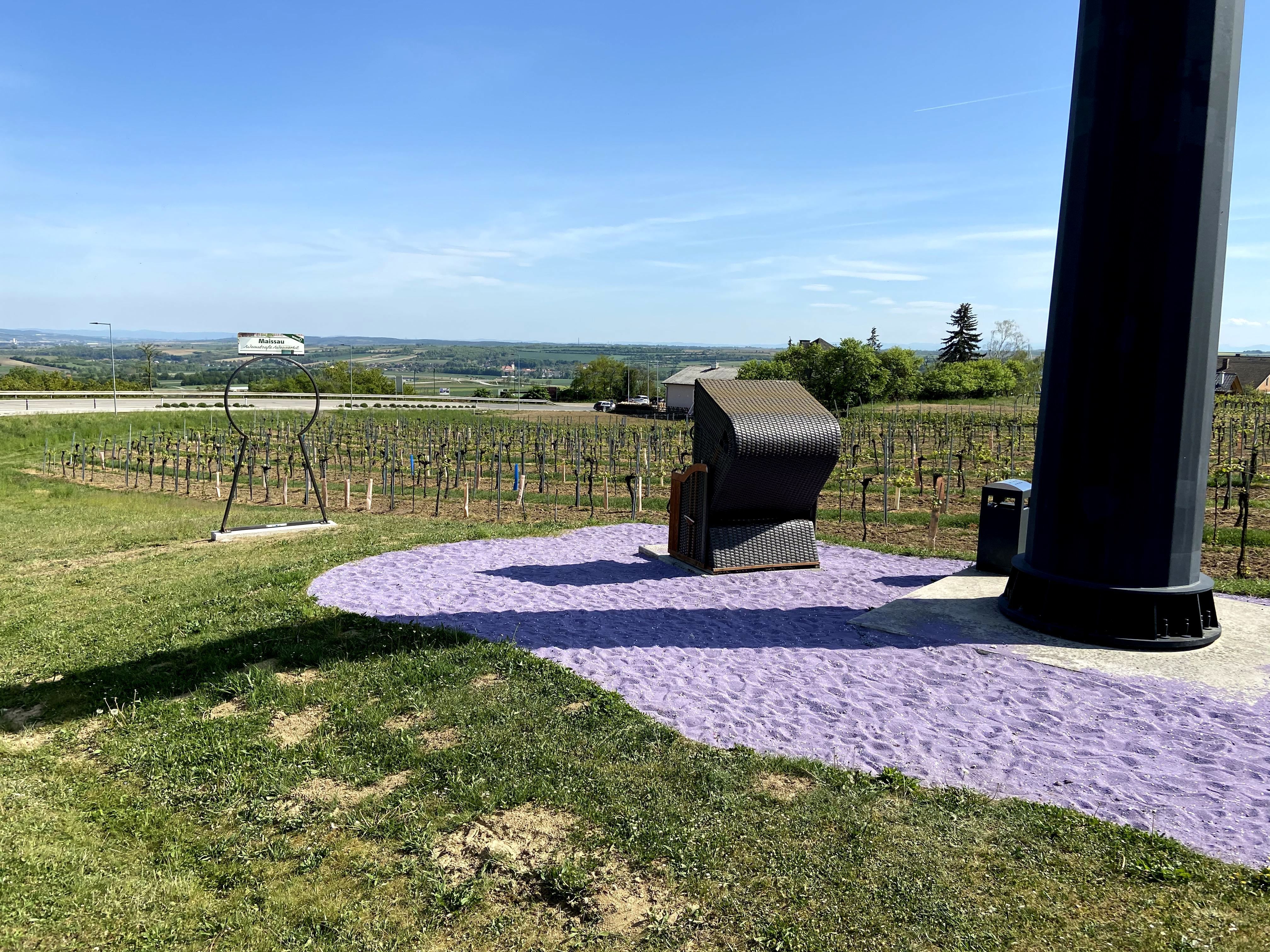 Ein Strandkorb steht auf violettem Sand neben einem großen schwarzen Pfeiler, umgeben von Weinbergen und einer weiten Landschaft.
