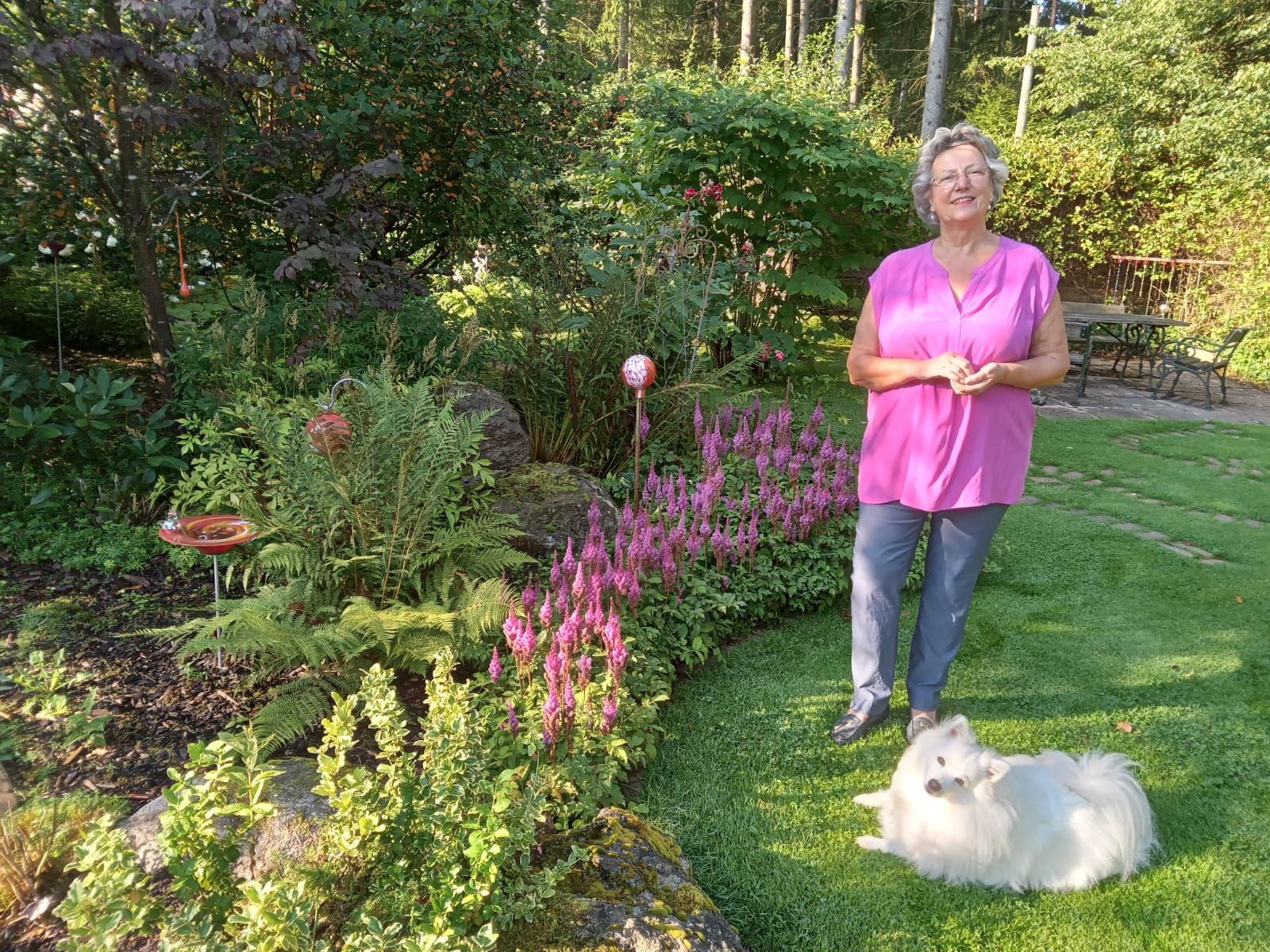 Eine Frau steht in einem Garten neben einem weißen Hund auf dem Rasen.