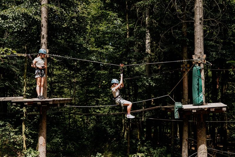 Zwei Kinder mit Helmen und Sicherheitsgurten in einem Kletterpark im Wald.