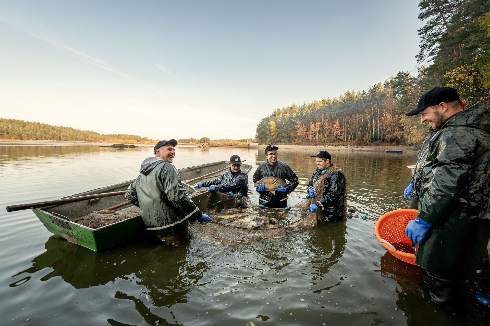 Ein fröhliches Team von Fischern steht am Ufer eines malerischen Teiches, umgeben von der ruhigen Natur. Die Sonne strahlt auf das Wasser und lässt die Fische im Netz glitzern, während die frische Luft den Duft von Abenteuer und Gemeinschaft verbreitet.