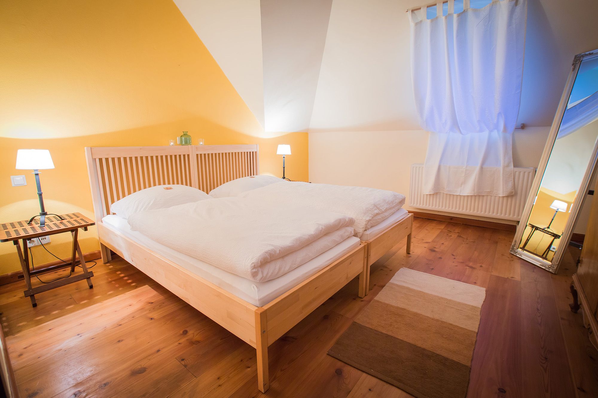 Gemütliches Schlafzimmer mit Doppelbett, Holzboden, gelber Wand und großem Spiegel.