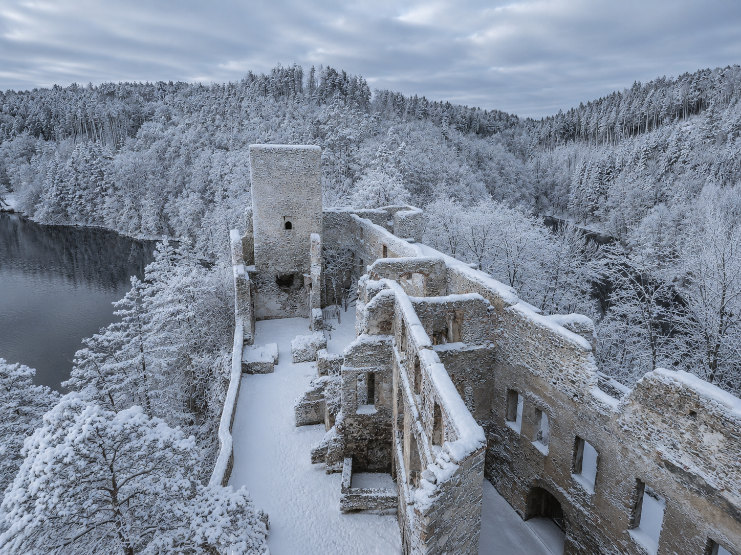 Ruine Dobra im Winter: verschneite Burgruine über einem Fluss, umgeben von dichtem Wald und bewaldeten Hügeln.