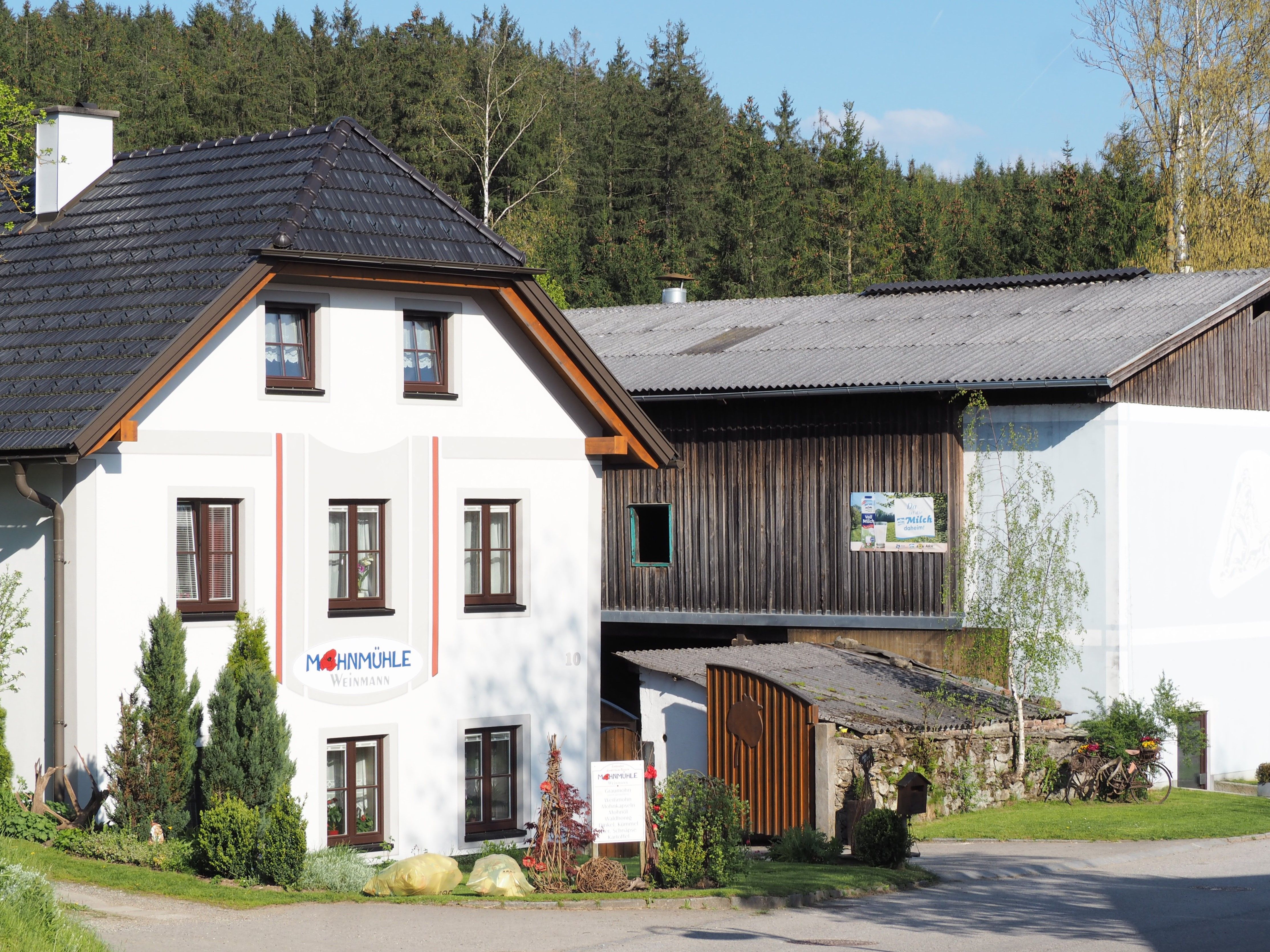 Gebäude der Mohnmühle Weinmann mit angrenzendem Holzschuppen, umgeben von Bäumen.