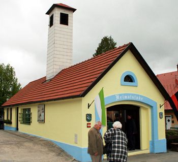 Kleines gelbes Gebäude mit rotem Dach und Schornstein, beschriftet mit 'Heimatstube'. Zwei Personen stehen davor.