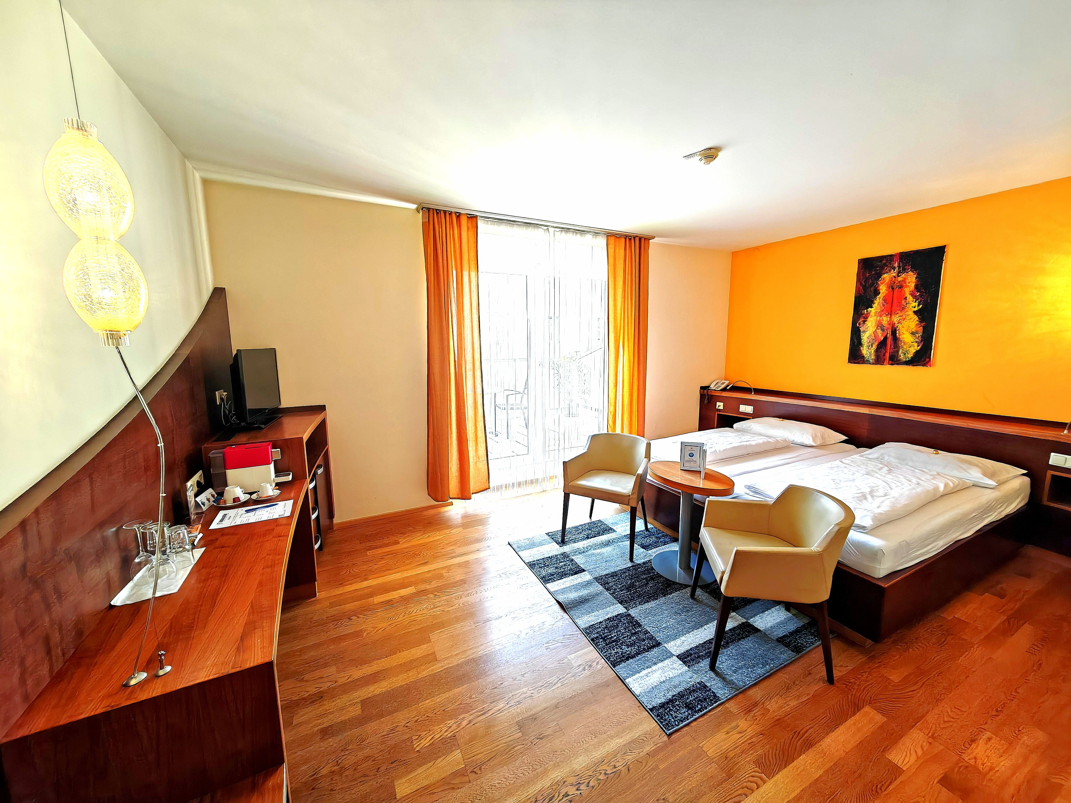 Ein modernes Hotelzimmer mit einem Doppelbett, zwei Sesseln, einem kleinen Tisch und einem Schreibtisch mit Fernseher. Die Wände sind in warmen Farben gestrichen.