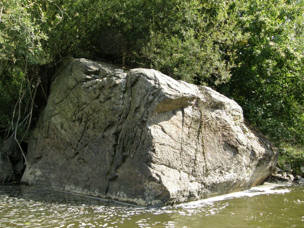 Großer Felsen am Ufer eines Flusses, umgeben von Bäumen.
