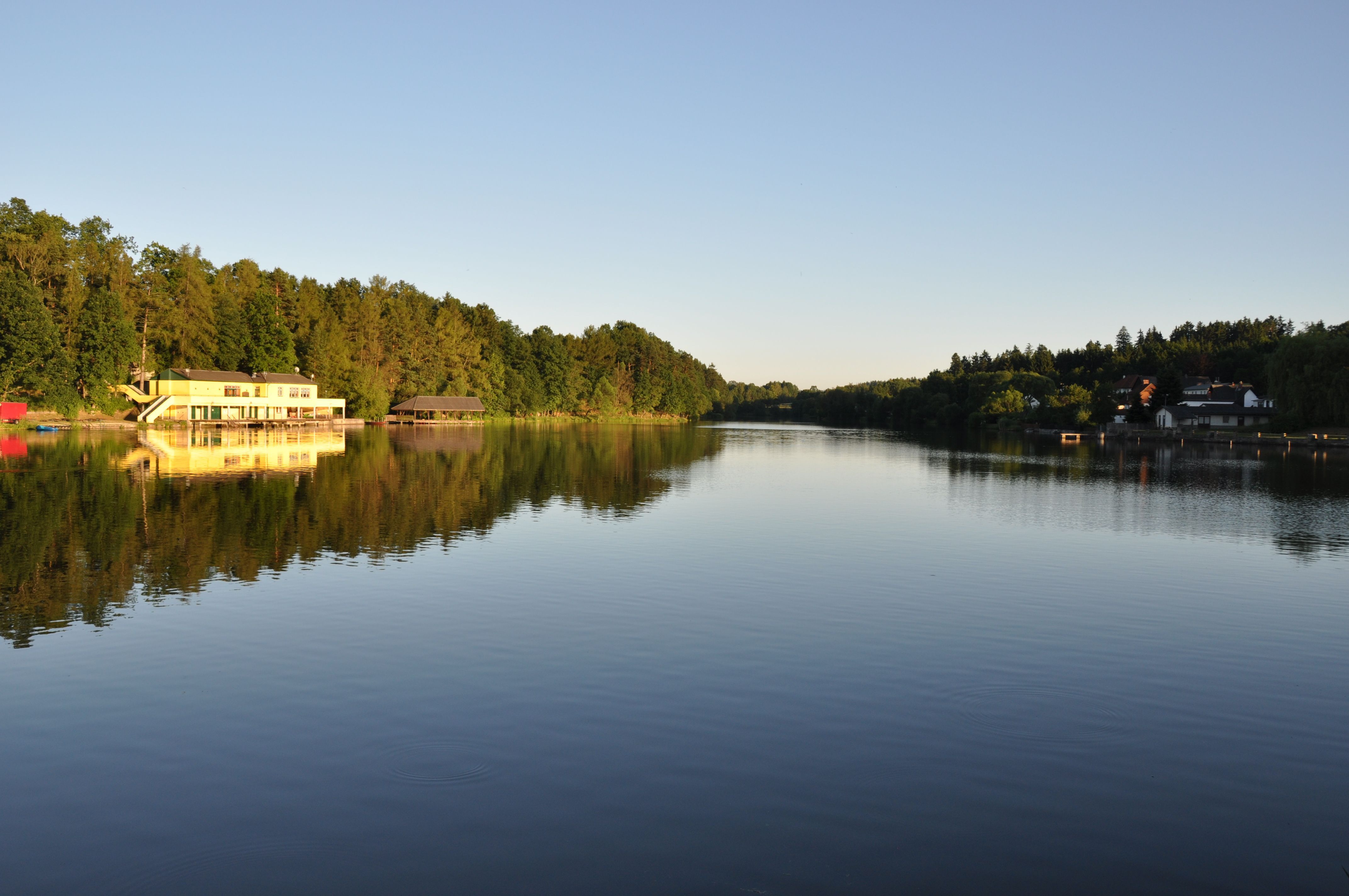 Ein ruhiger See mit einem Gebäude am Ufer, umgeben von Bäumen, bei Sonnenuntergang.