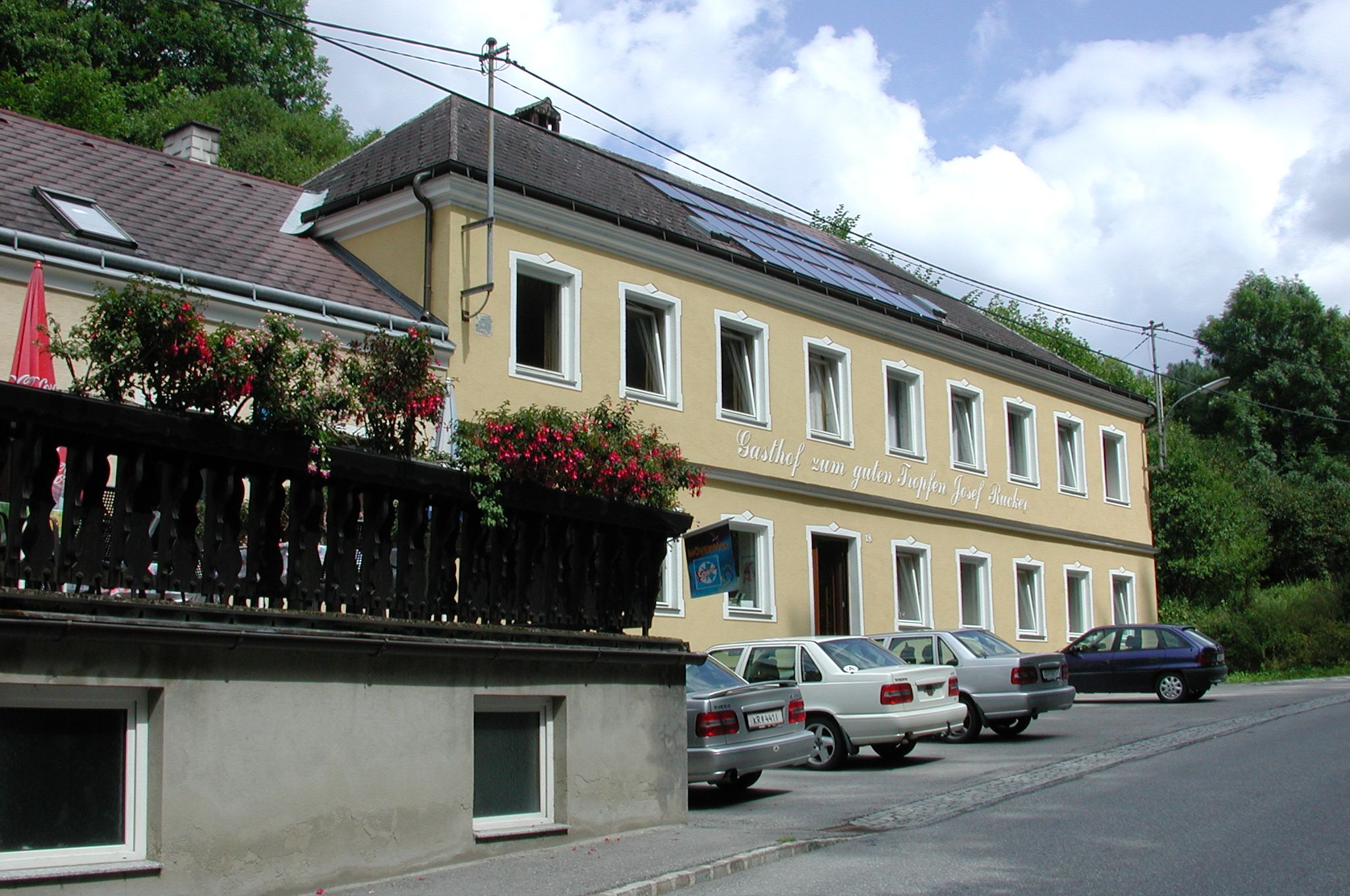 Gelbes Gebäude mit der Aufschrift 'Gasthof zum guten Tropfen Josef Rucker', Autos parken davor, blauer Himmel im Hintergrund.