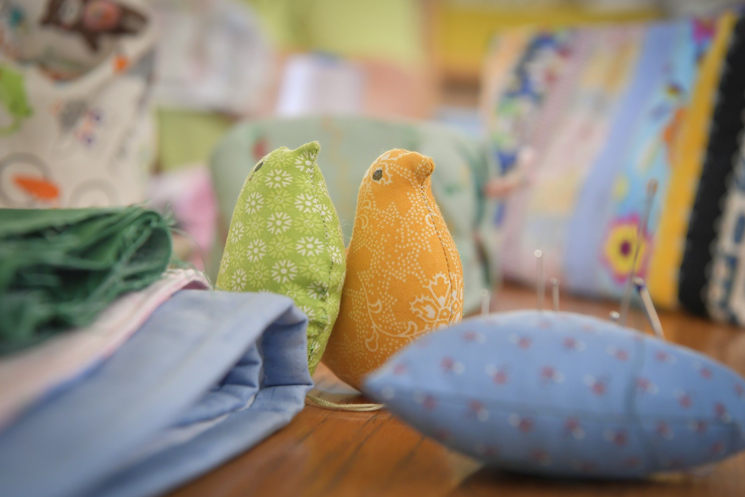 Zwei Stoffvögel, einer grün und einer orange, auf einem Tisch mit Stoffen und Nähutensilien.