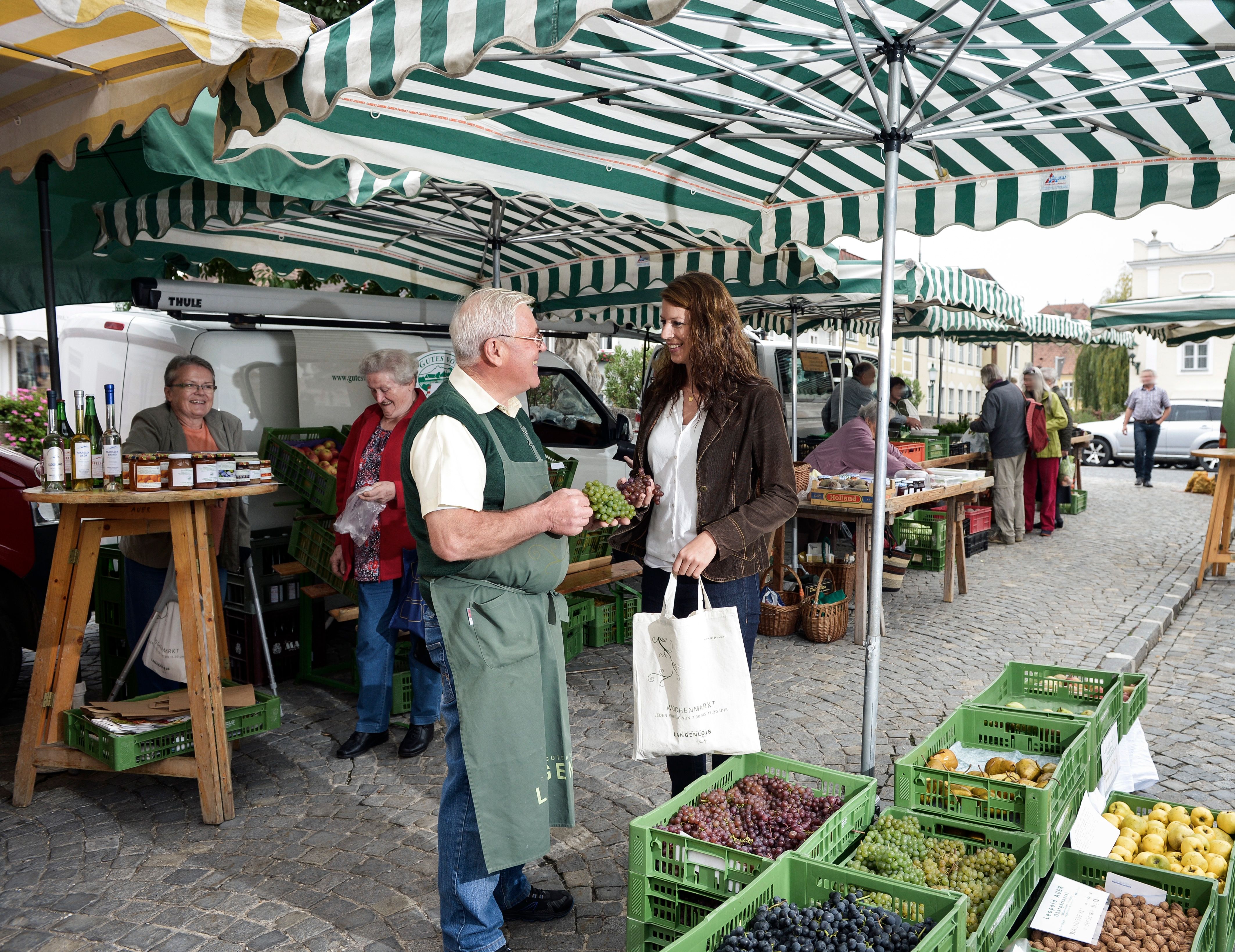 Menschen auf einem Wochenmarkt mit Obst- und Gemüseständen unter gestreiften Markisen.