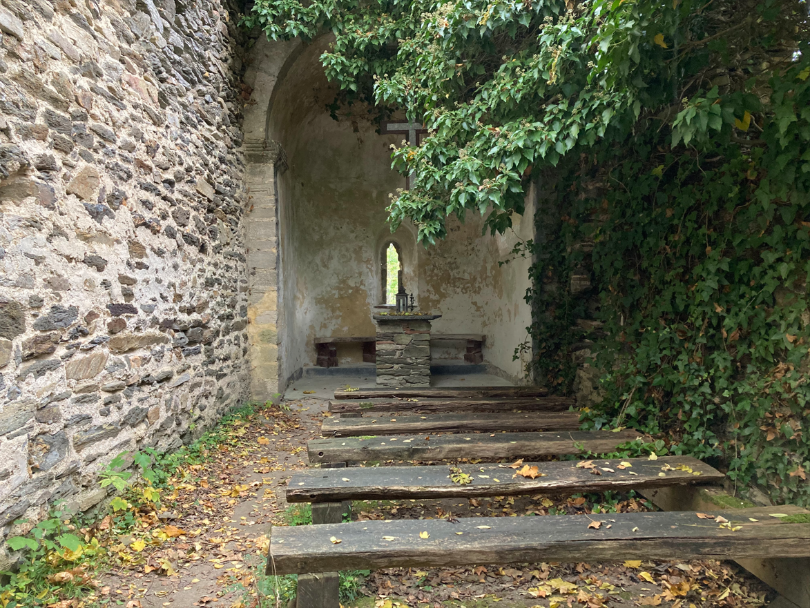 Verlassene Kapelle mit Steinmauern und Holzbänken, umgeben von Efeu.