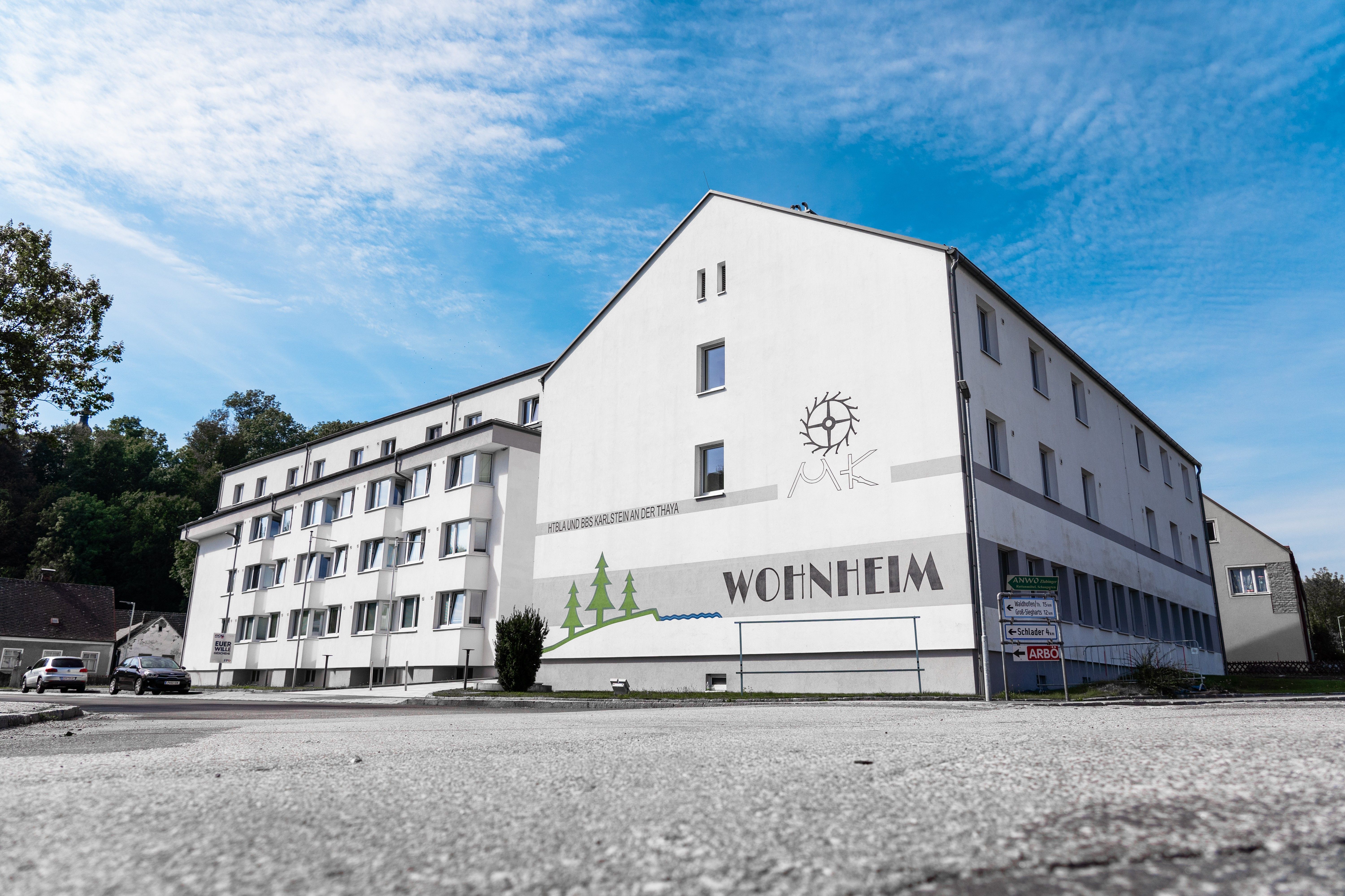 Ein großes, weißes Gebäude mit der Aufschrift 'Wohnheim' und einem Logo an der Wand. Der Himmel ist blau mit einigen Wolken.