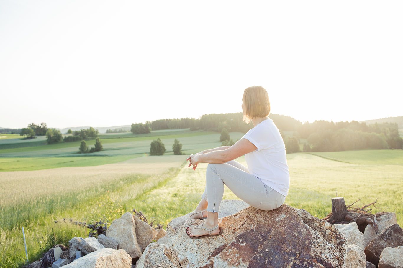 Person sitzt auf einem Felsen und blickt über eine grüne Landschaft im Sonnenlicht.