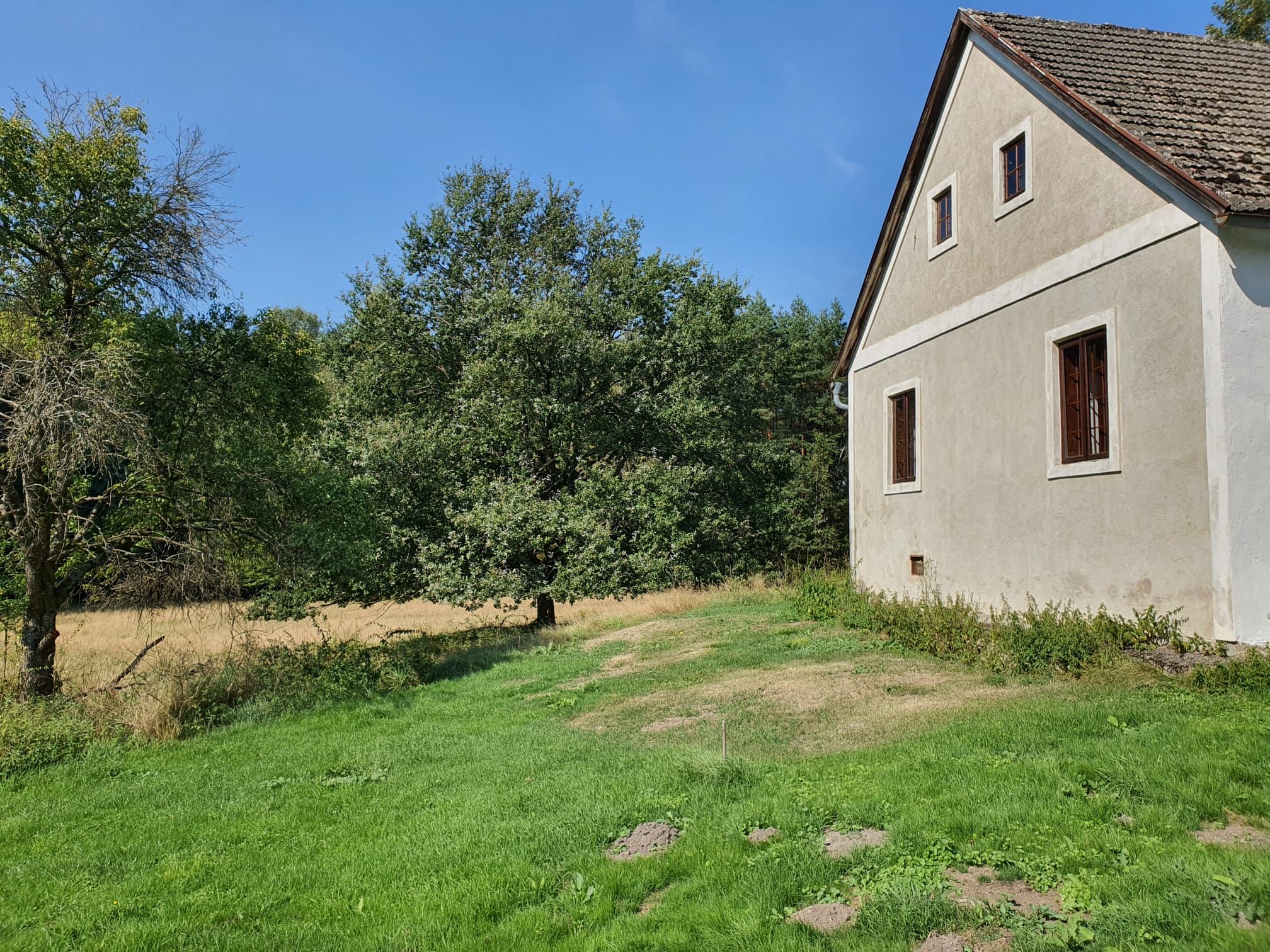 Ein altes Haus mit braunem Dach und weißen Wänden steht neben einem grünen Rasen und Bäumen unter einem klaren blauen Himmel.