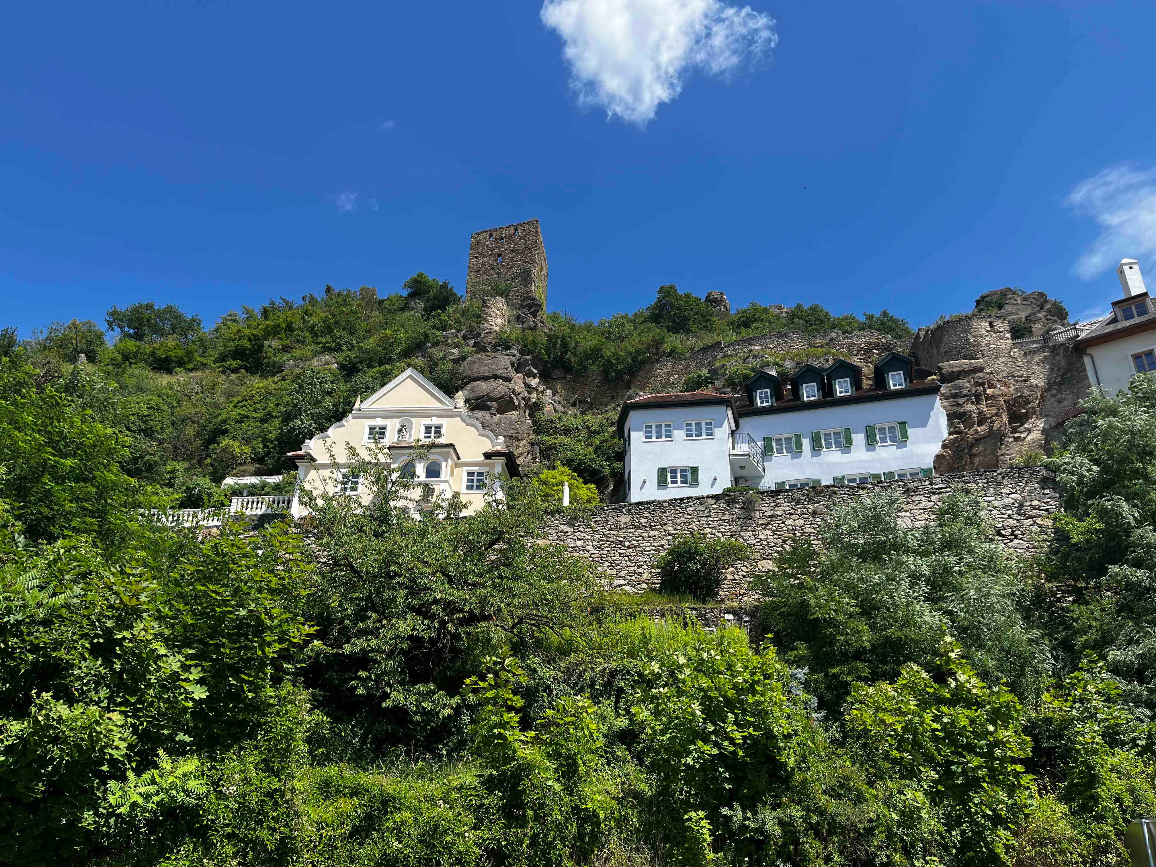 Blick auf Dürnstein mit historischen Gebäuden und Ruinen auf einem Hügel, umgeben von grüner Vegetation und blauem Himmel.