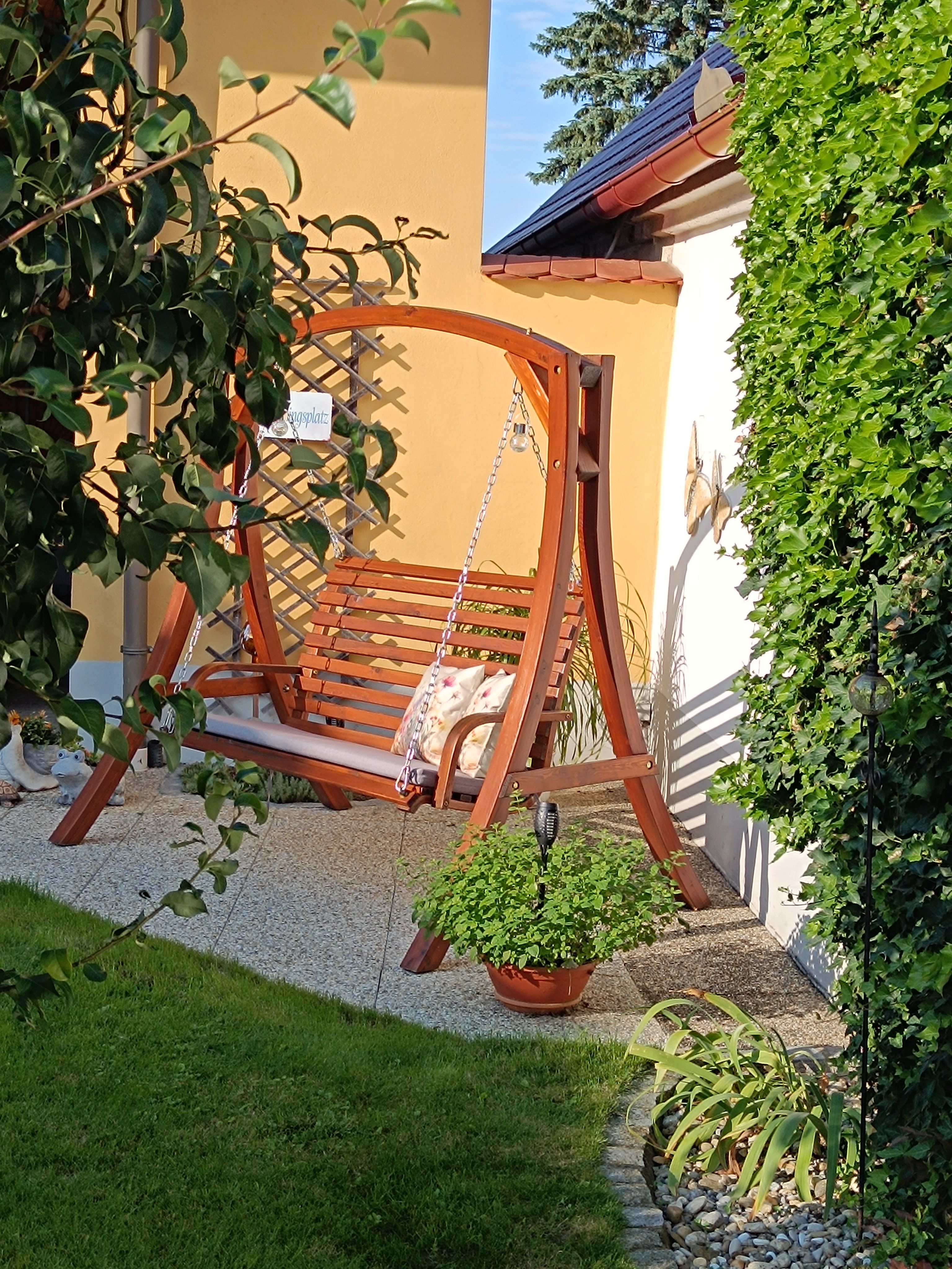 Holzschaukel im sonnigen Innenhof mit Pflanzen und grüner Hecke.