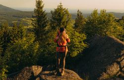 Ein Wanderer steht auf einem Felsen mit Ausblick auf bewaldete Hügel, aufgenommen im Waldviertel, gekrönt von einem klaren Himmel und dem Licht der aufgehenden Sonne.