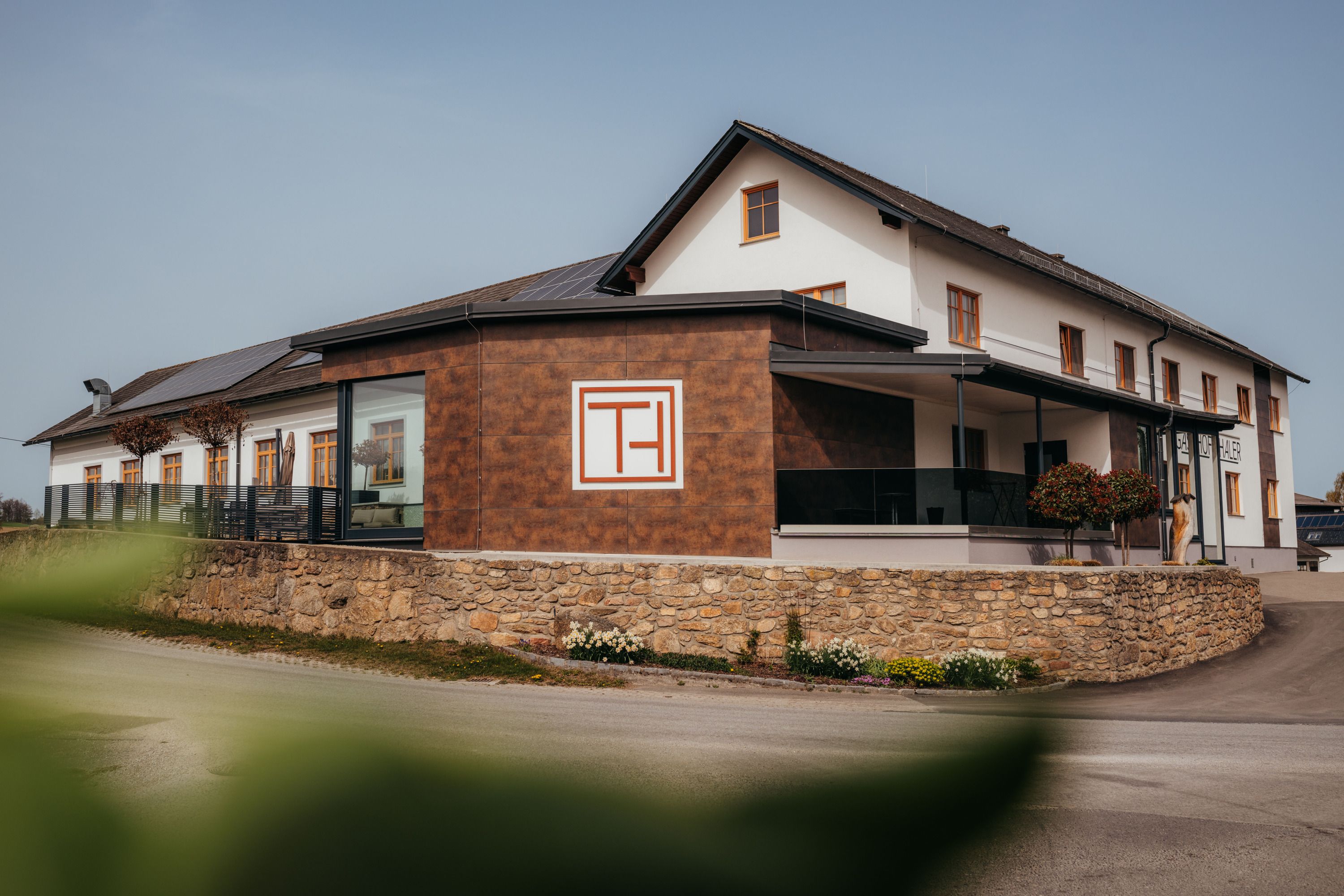 Ein modernes Gebäude mit der Aufschrift 'Gasthof Thaler', umgeben von einer Steinmauer und Pflanzen.