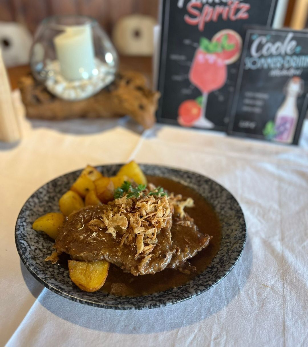 Ein Teller mit Bratkartoffeln und einem Stück Rindfleisch, garniert mit Röstzwiebeln, auf einem Tisch mit Dekoration und Getränkekarten im Hintergrund.