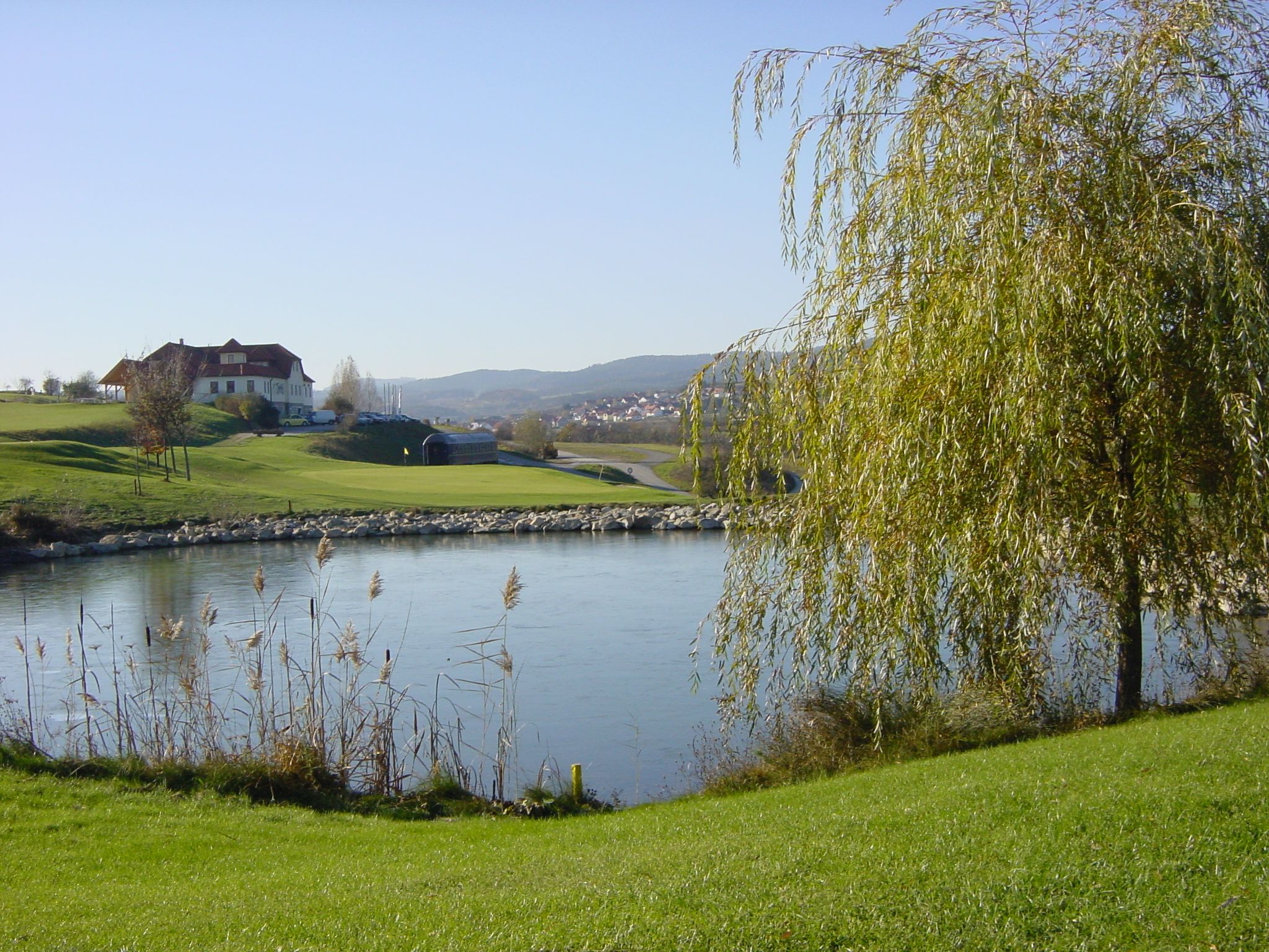 Landschaft mit Golfplatz, Teich und Gebäude im Hintergrund.