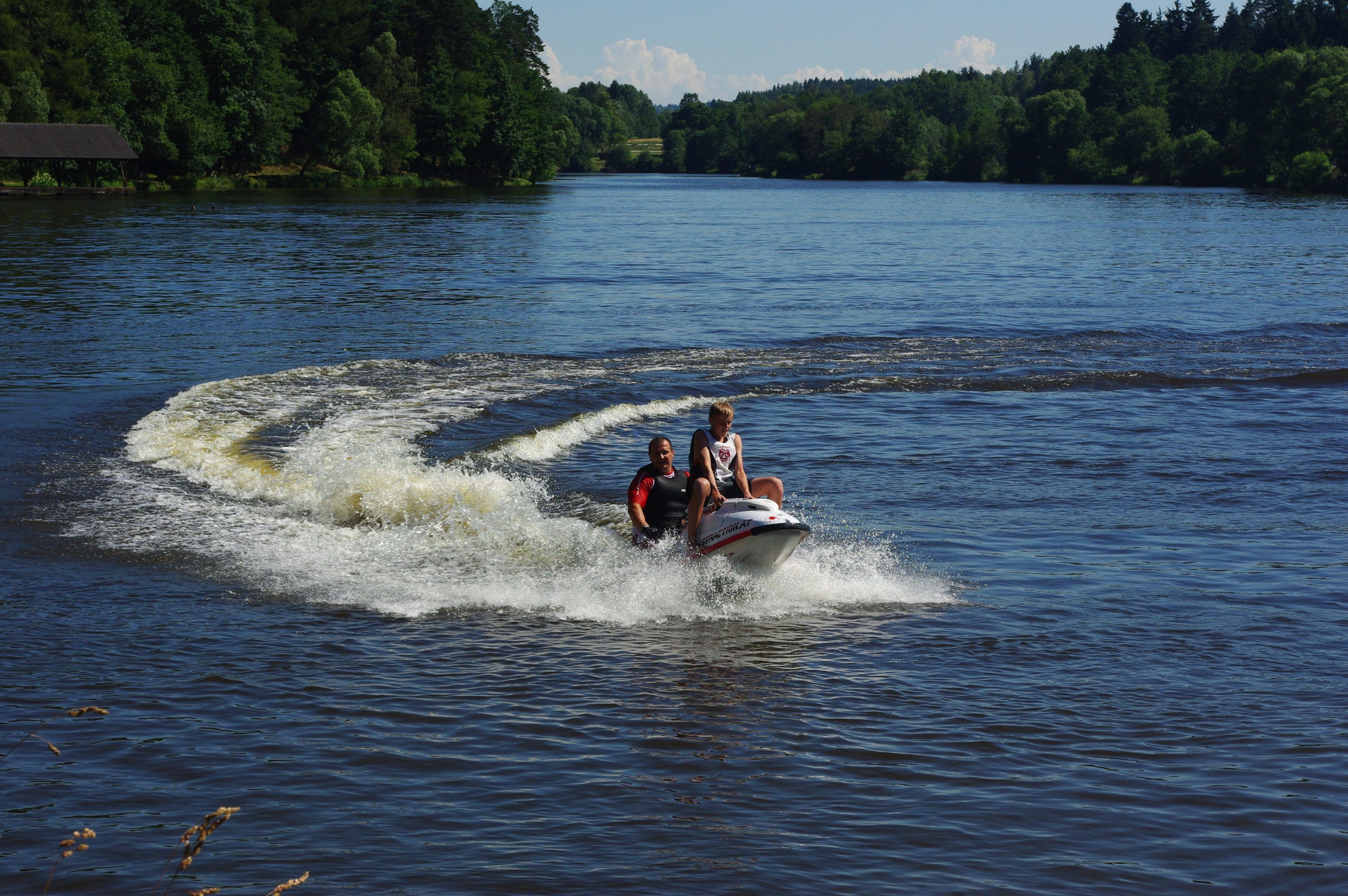 Zwei Personen fahren auf einem Jetski über einen See, umgeben von Bäumen.