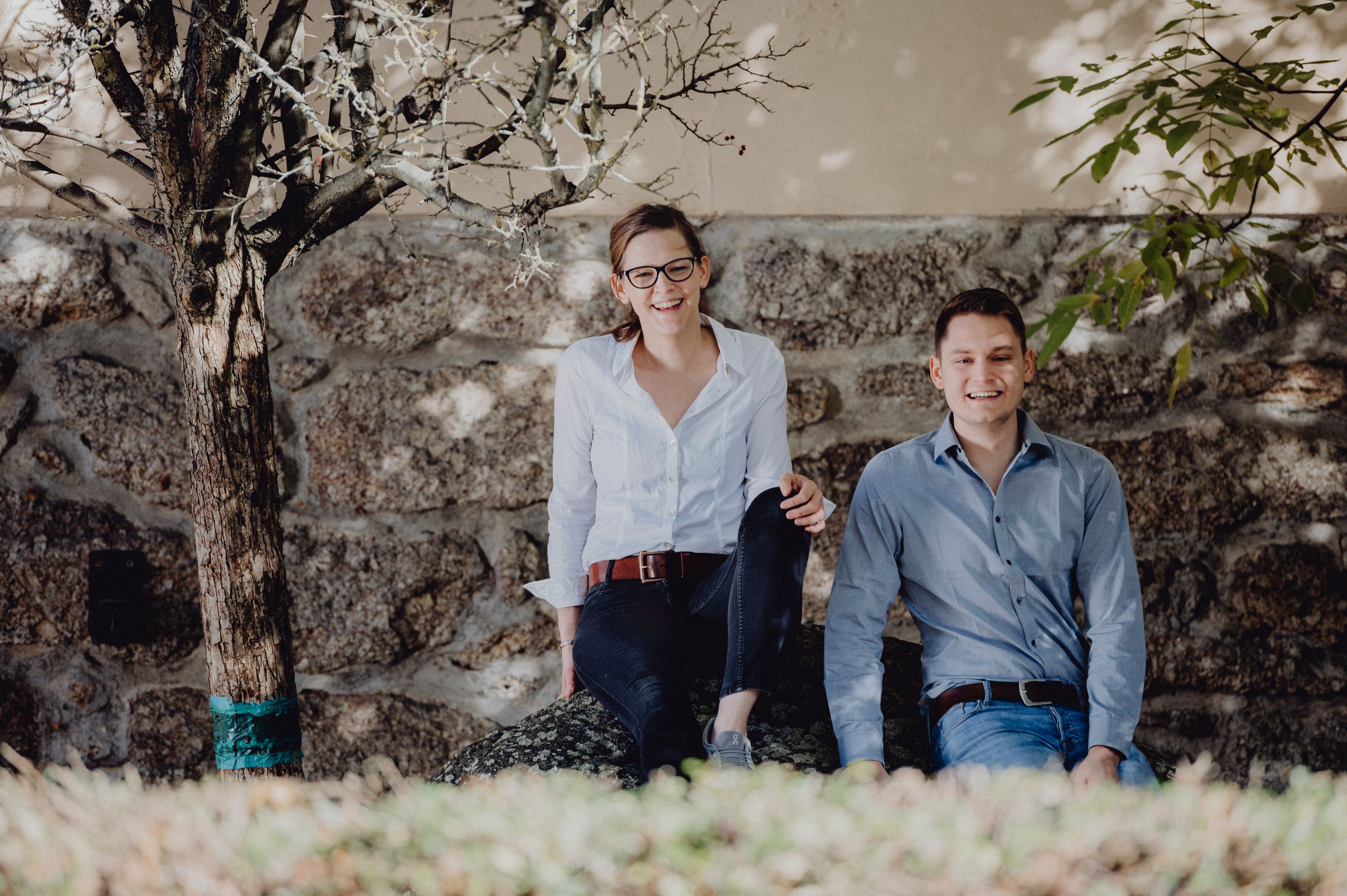 Zwei Personen sitzen lächelnd auf einem Stein vor einer Steinmauer, umgeben von Bäumen.