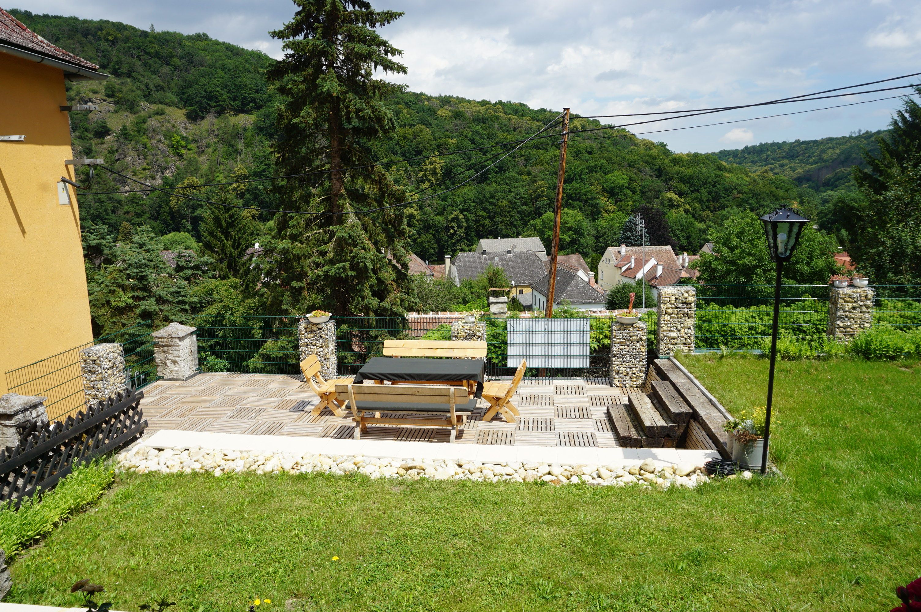 Terrasse mit Holzmöbeln und Blick auf ein Dorf und bewaldete Hügel.