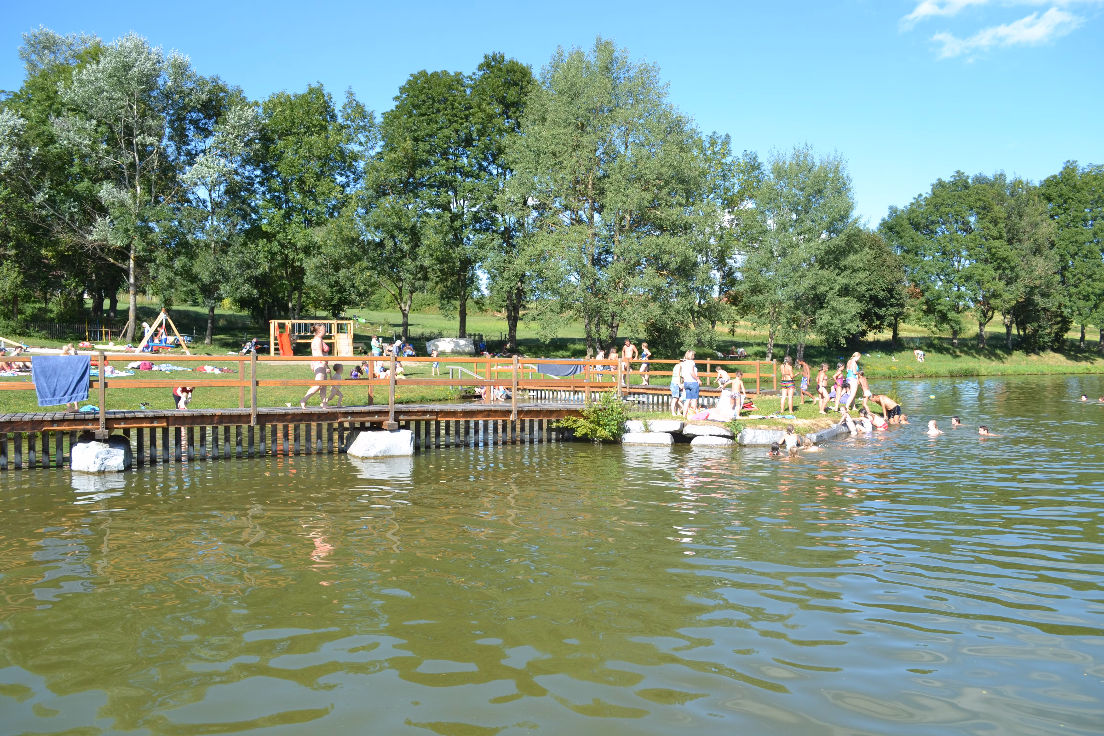 Menschen schwimmen und entspannen an einem Badeteich mit Steg und Spielplatz im Hintergrund.