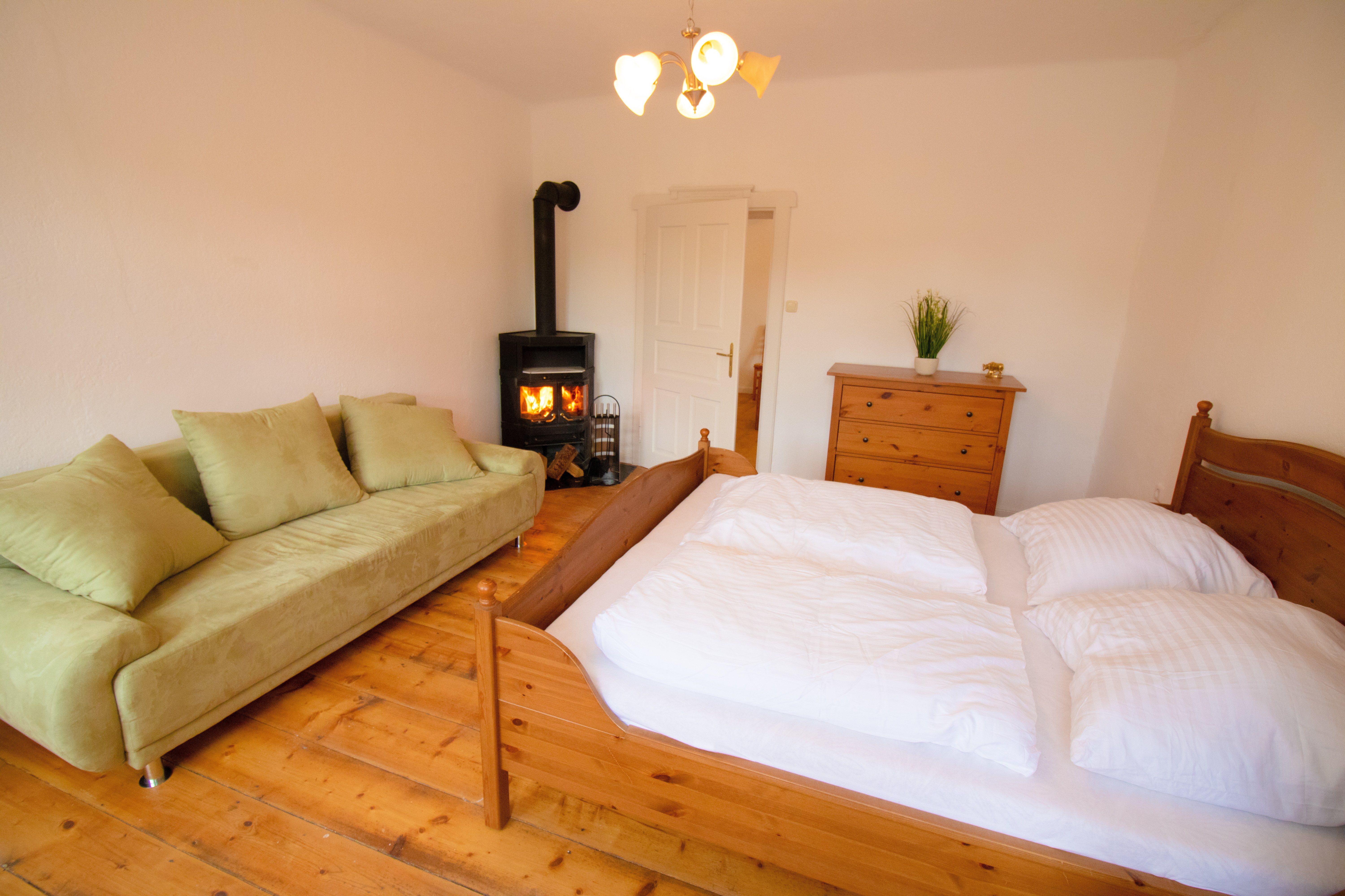 Gemütliches Schlafzimmer mit Holzfußboden, grünem Sofa, Holzofen, Doppelbett und Kommode.