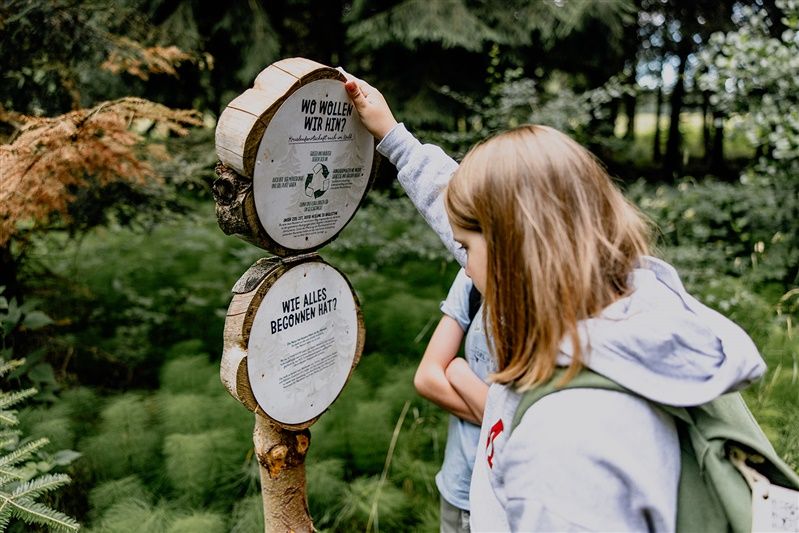 Ein Mädchen betrachtet ein hölzernes Schild im Wald mit Informationen über die Natur.