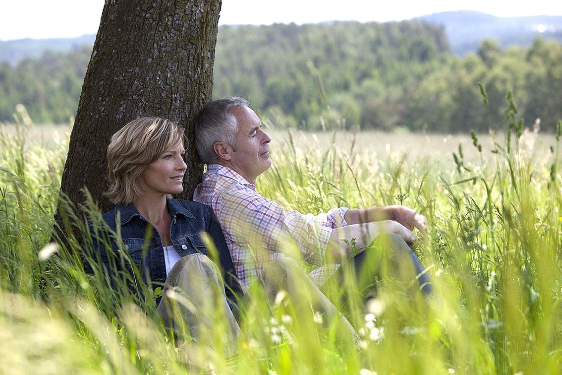 Ein Paar sitzt entspannt an einen Baum gelehnt in einer grünen Wiese.