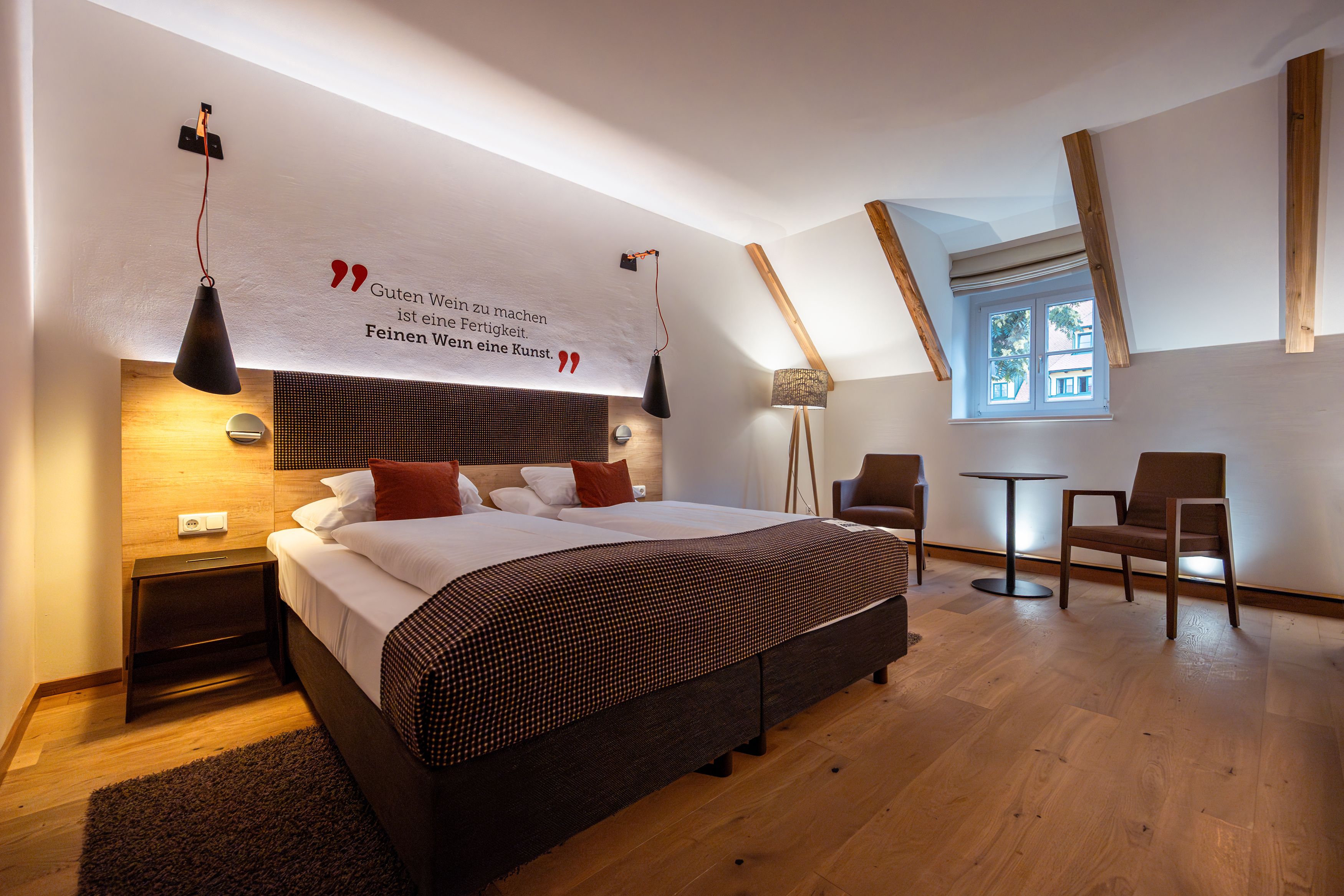 Ein stilvoll eingerichtetes Hotelzimmer mit Doppelbett, zwei Sesseln und einem kleinen Tisch. An der Wand steht ein Zitat über Wein.