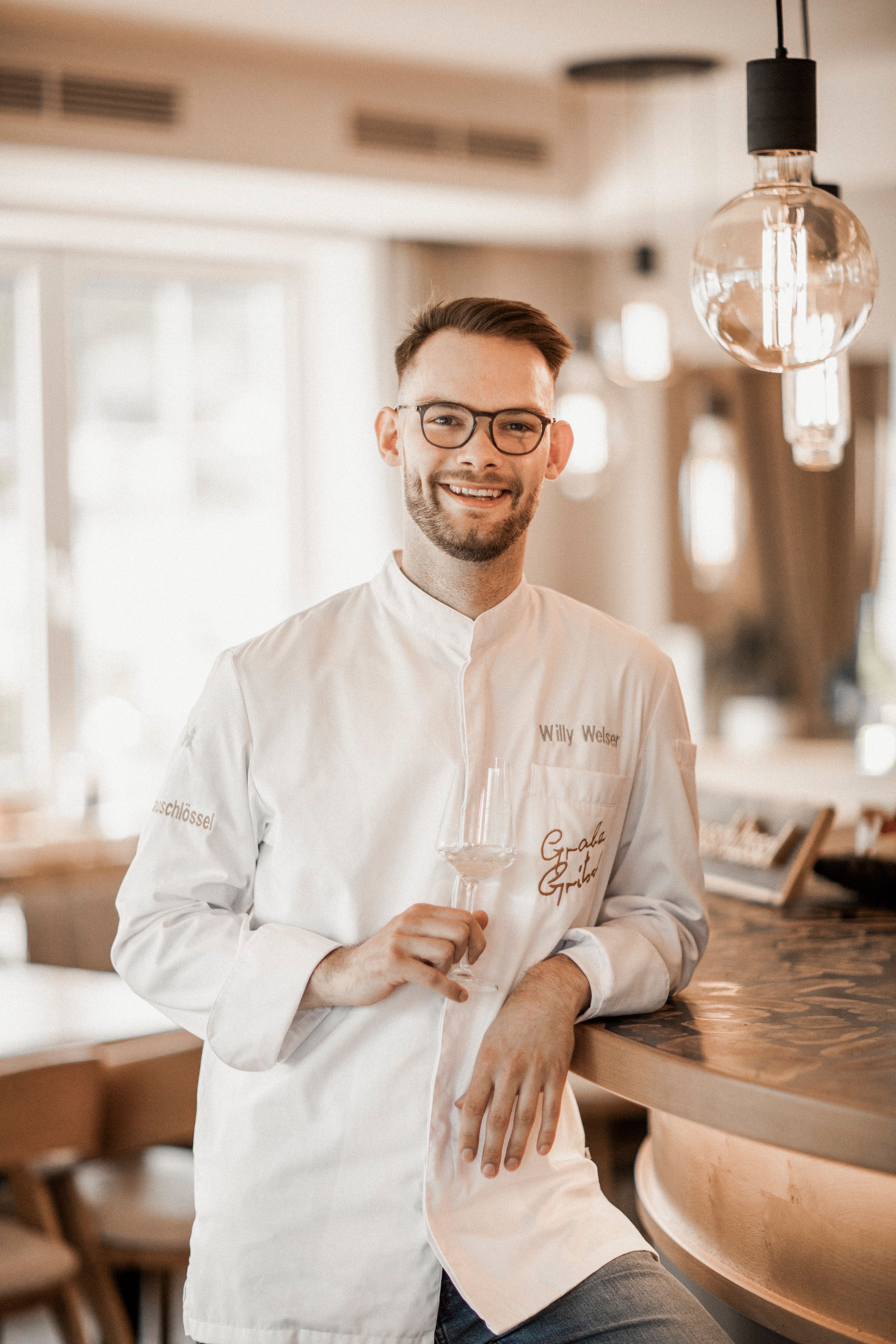 Ein lächelnder Mann in Kochjacke steht an einer Bar und hält ein Weinglas.