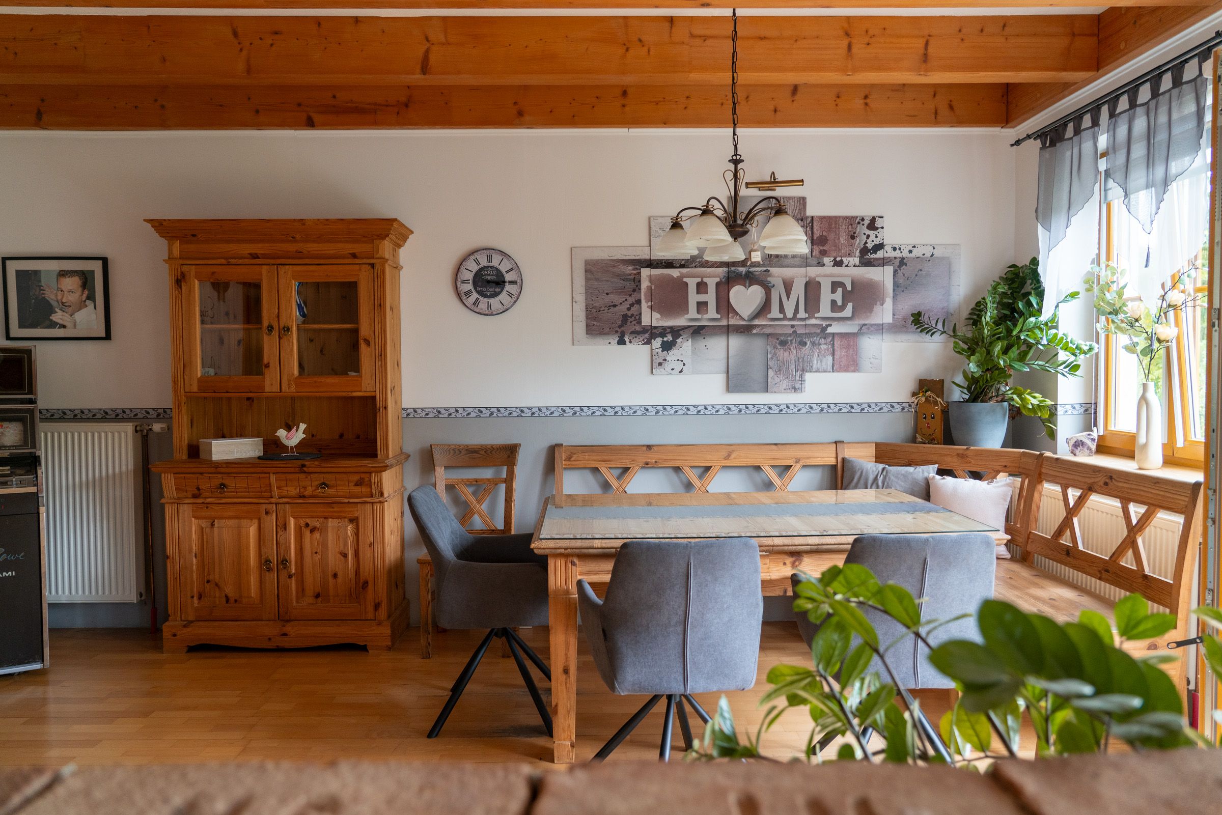 Gemütliche Küche mit Holzmöbeln, Esstisch, grauen Stühlen und Pflanzen. An der Wand ein "Home"-Schild und eine Uhr.