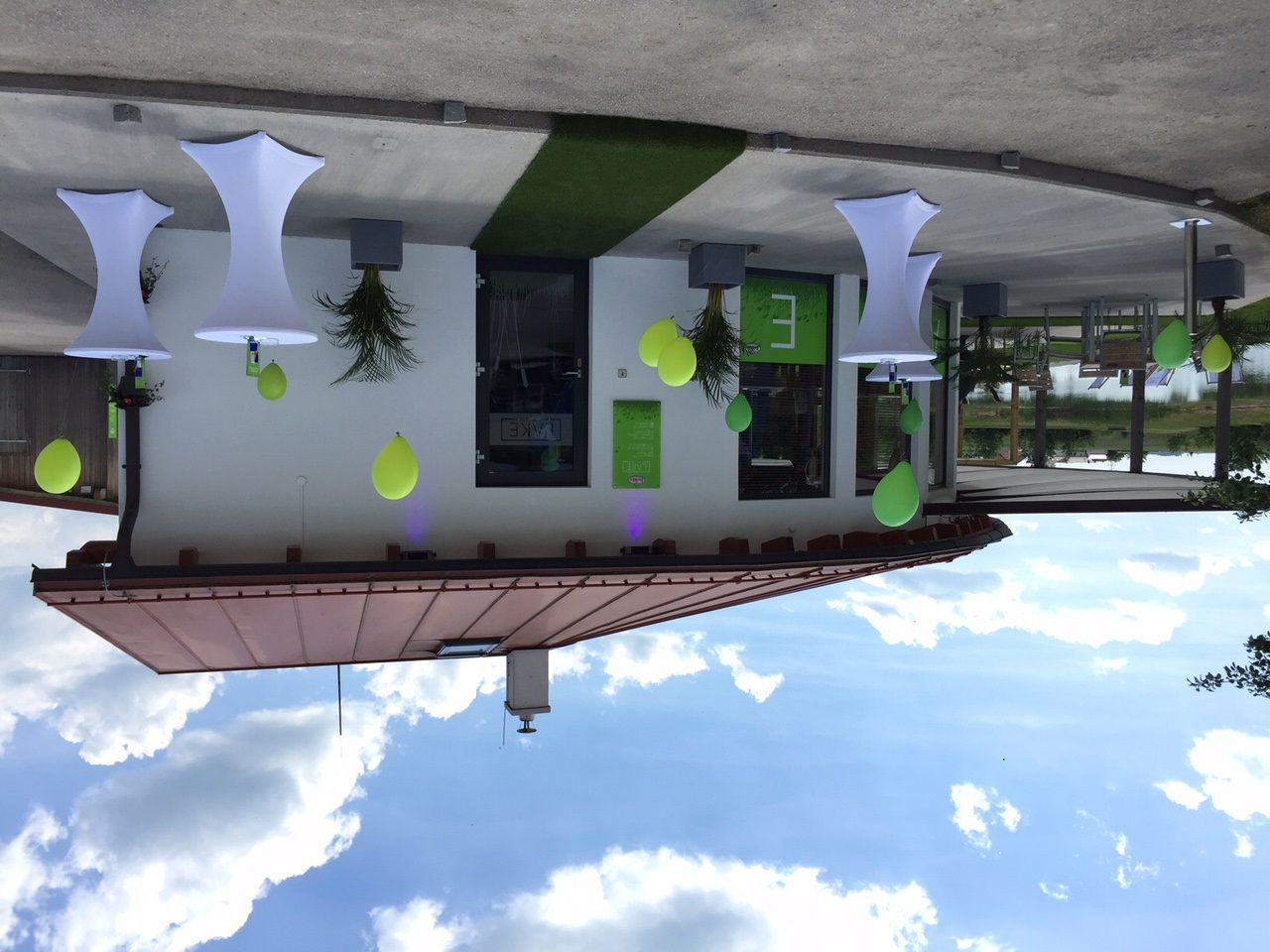 Eine moderne Strandbar mit weißen Stehtischen und grünen Dekorationen vor einem Gebäude mit Flachdach.