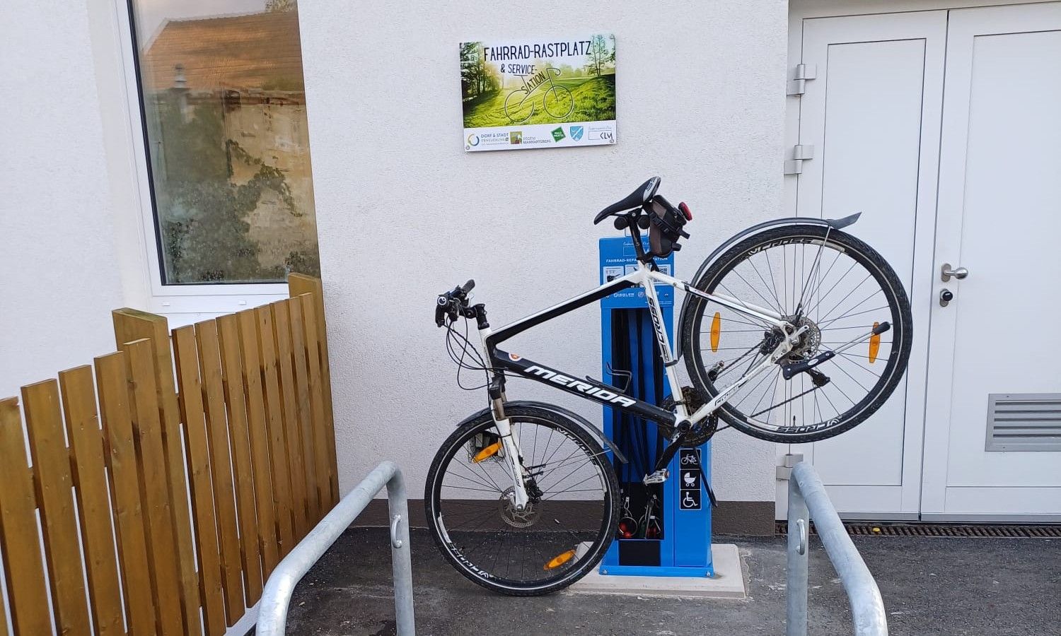 Fahrrad an einer blauen Service-Station befestigt, neben einem Gebäude mit einem Schild 'Fahrrad-Rastplatz & Service Station'.