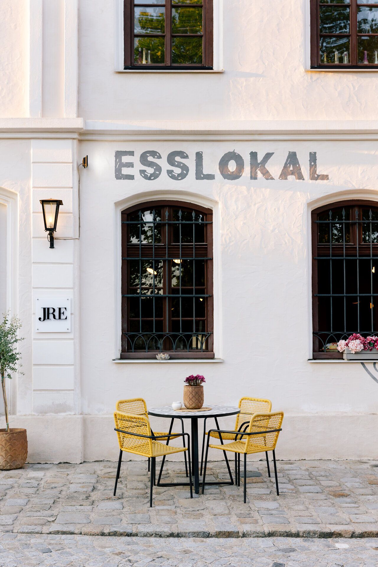 Vor einem charmanten Esslokal laden gelbe Stühle und ein eleganter Tisch dazu ein, die frische Luft und die köstlichen Aromen der Region zu genießen. Die sanften Farben der Fassade harmonieren perfekt mit der blühenden Umgebung und schaffen eine einladende Atmosphäre für alle, die eine Auszeit suchen. Hier wird der Genuss zum Erlebnis, während man die Schönheit der Natur in vollen Zügen auskostet.